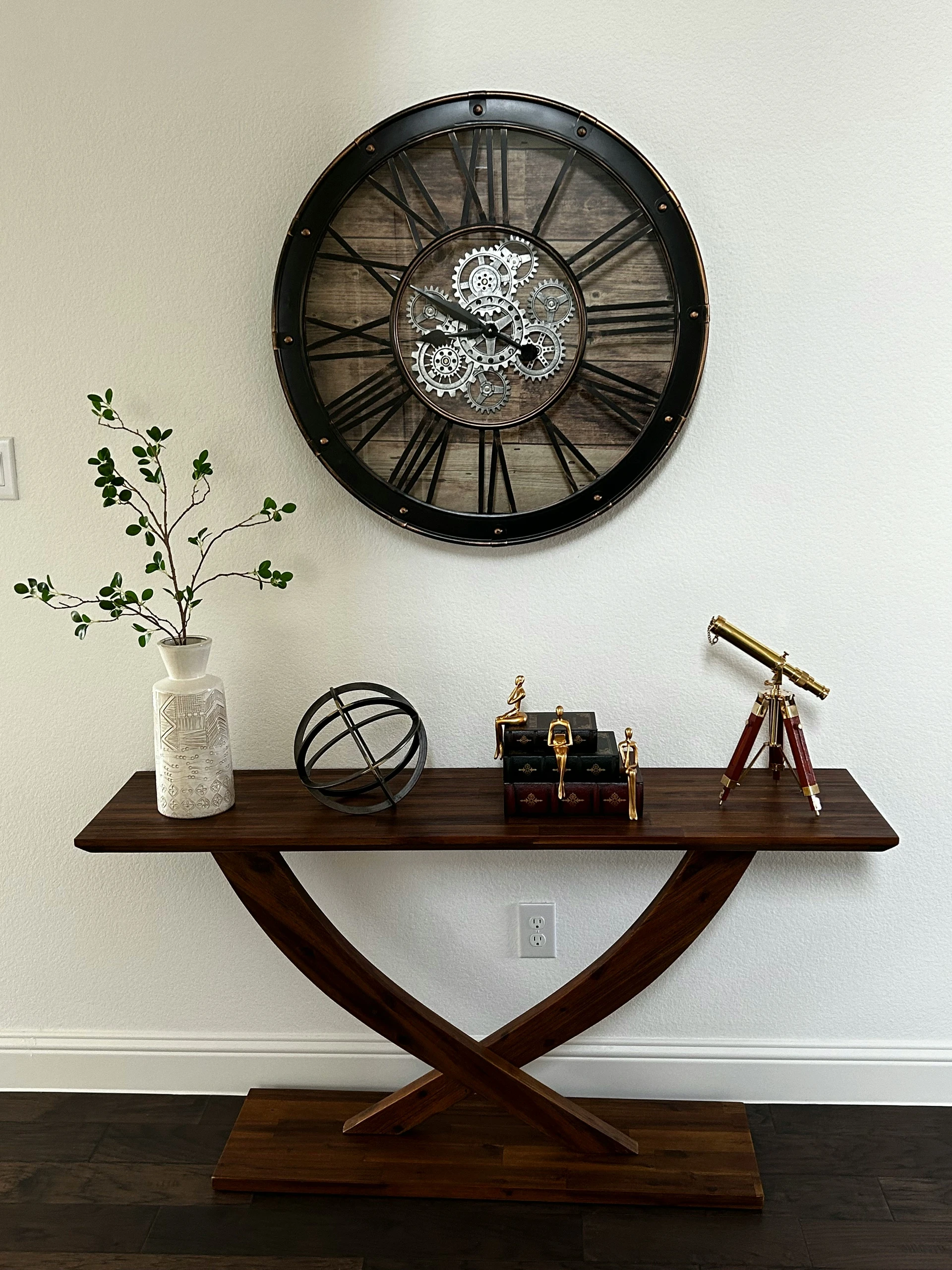 Elegant living room vignette featuring a sculptural wooden console table adorned with decorative items like a ceramic vase, metal sphere, miniature figurines, and a brass telescope. Above the table hangs a striking artistic clock with exposed gears and Roman numerals, set against a rustic wood backdrop. A refined example of living room decor ideas for walls, blending industrial charm with sophisticated accents.