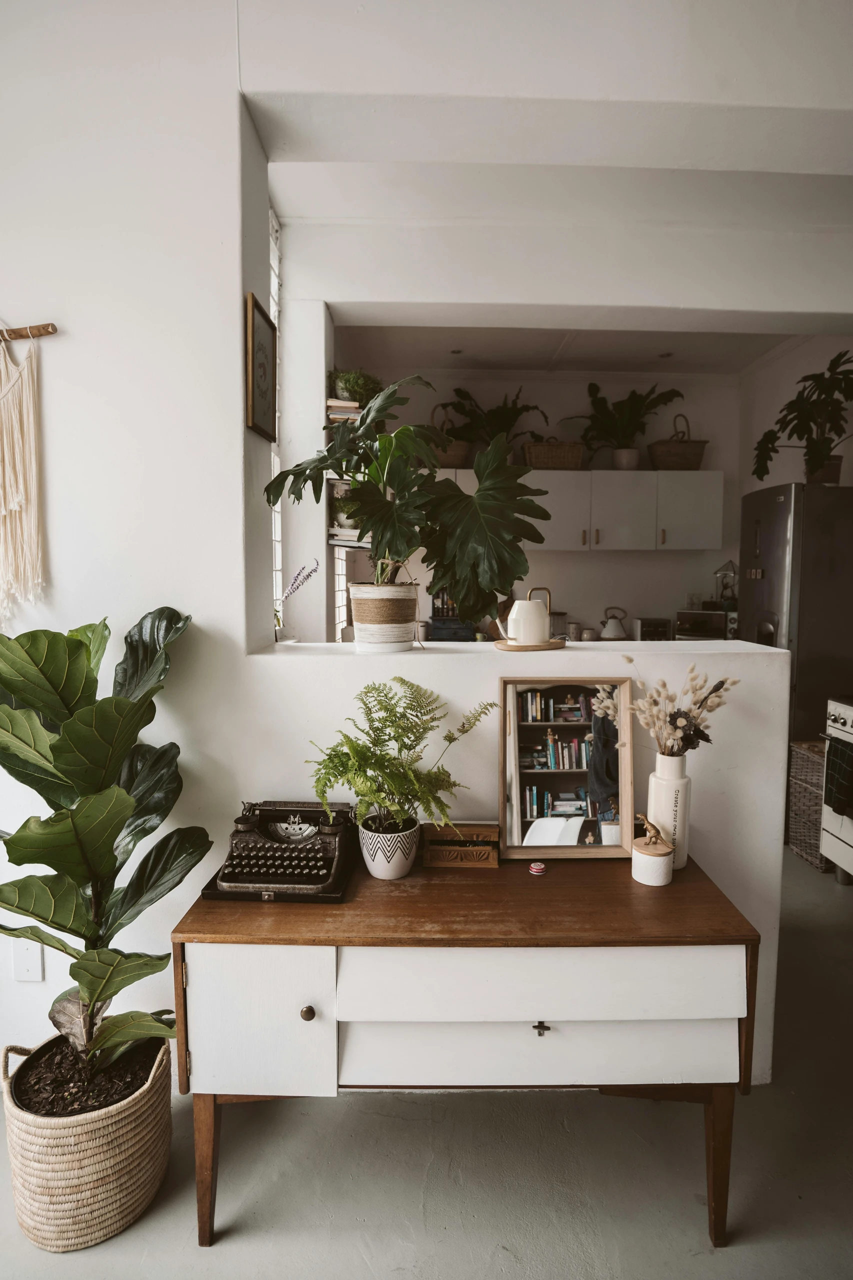 Stylish living room corner featuring a mid-century modern console table decorated with potted plants, a vintage typewriter, and a framed mirror, surrounded by greenery and soft lighting. Perfect inspiration for cozy and functional living room decor ideas for walls.