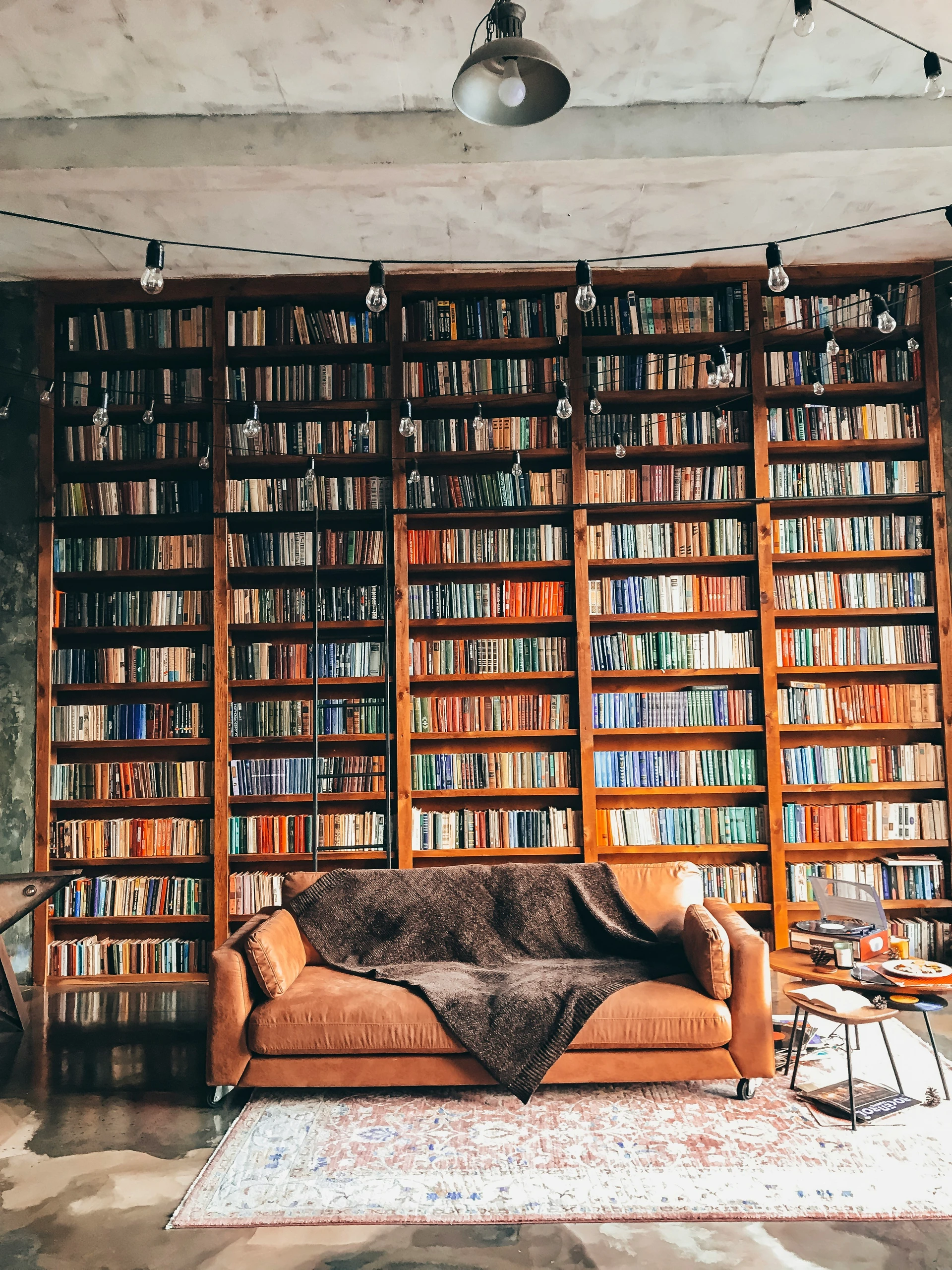 Cozy and eclectic living room featuring a warm-toned leather sofa with a dark throw blanket, set against a striking huge bookshelf that spans the entire wall from floor to ceiling. The shelves are filled with colorful books, creating a vibrant and intellectual backdrop. String lights and a vintage rug add charm to the space, showcasing creative and functional living room decor ideas for walls that combine personality, warmth, and style.