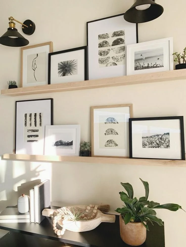 Modern and cozy living room corner featuring two light wood floating shelves displaying a curated collection of layered framed art work in black, white, and neutral tones. The prints include botanical sketches, abstract shapes, and serene landscapes, arranged in a visually balanced, overlapping style. Accented by matte black sconces, natural wood decor, and potted plants, this space offers stylish and versatile living room decor ideas for walls that blend minimalism with warmth.