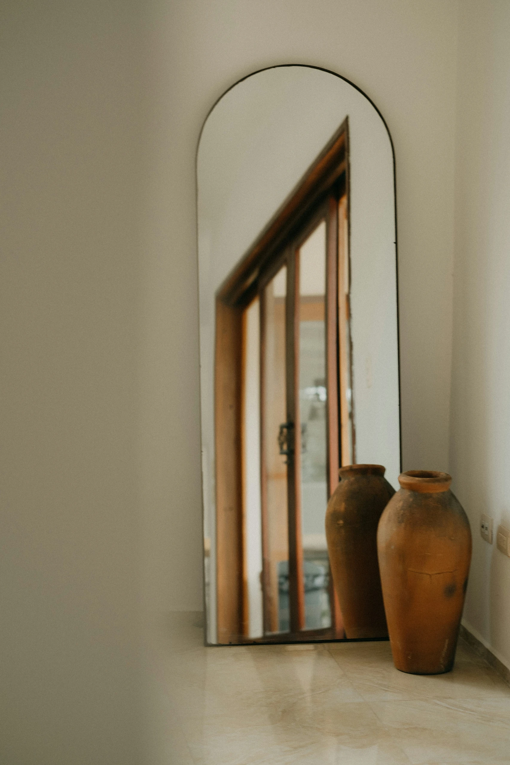 Minimalist living room corner featuring an oversized mirror with an arched top, leaning against a white wall. Two rustic terracotta vases add earthy texture and warmth to the neutral-toned space. A refined and functional example of living room decor ideas for walls, using reflective surfaces to enhance light and depth in modern interiors.