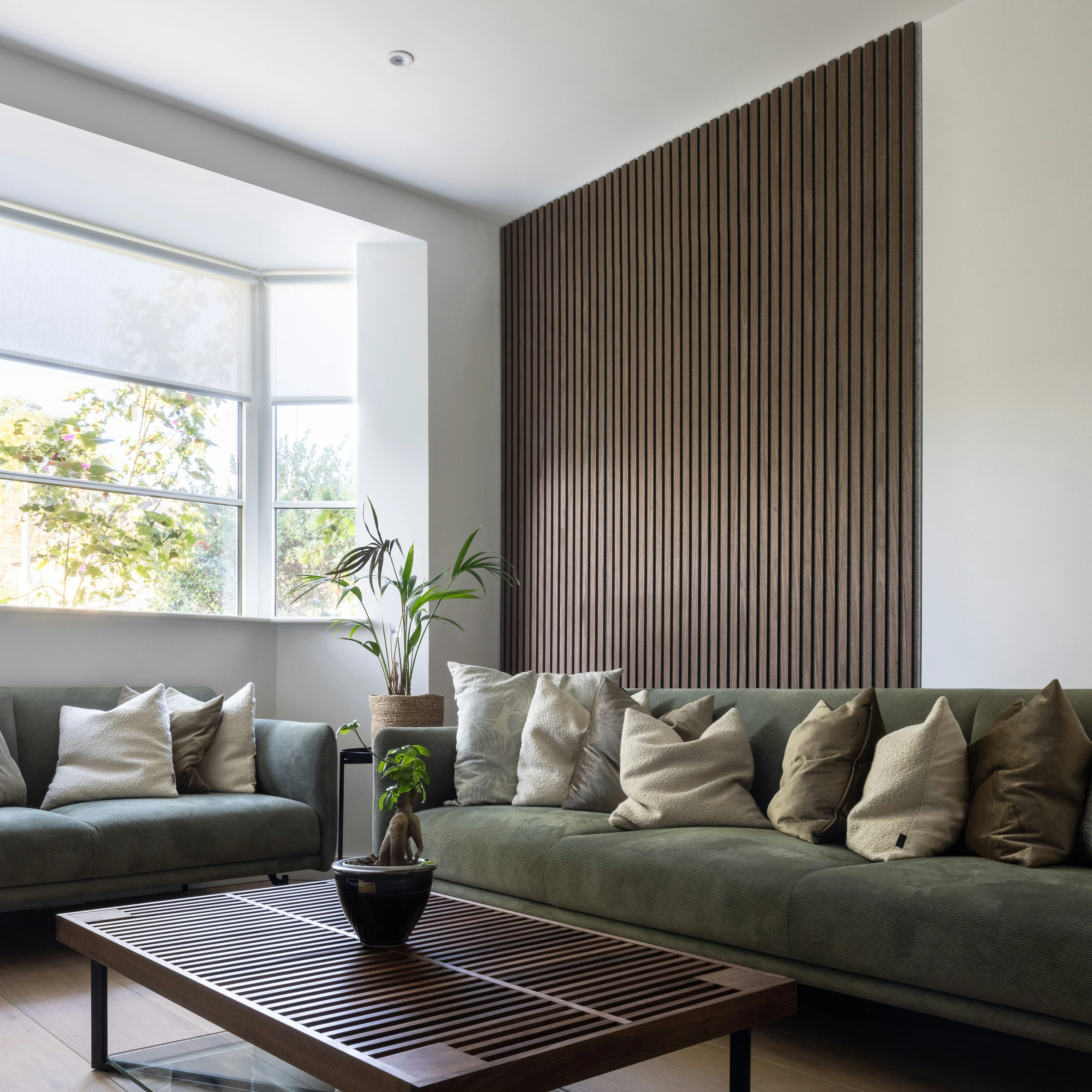 Modern living room with green velvet sectional sofas, neutral throw pillows, and a sleek wooden coffee table. The standout feature is the vertical wall paneling in a rich brown tone, adding texture and warmth to the minimalist design. Large windows bring in natural light, highlighting this stylish example of living room decor ideas for walls that combine functionality with contemporary elegance.