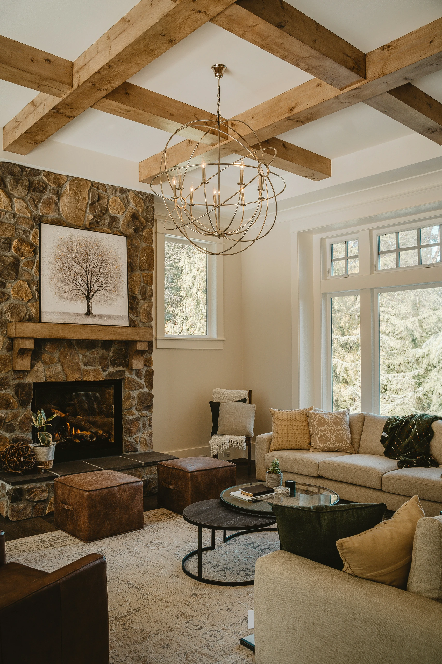 Cozy and elegant living room showcasing living room farmhouse decor ideas with a rustic stone fireplace, wooden ceiling beams, and various light sources including a large modern chandelier and natural light from expansive windows.