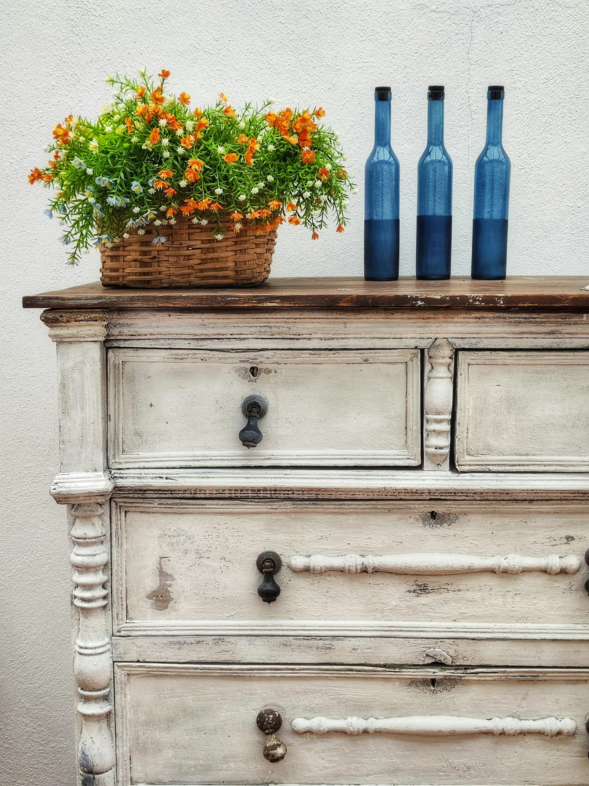 Alt text: A distressed white vintage dresser topped with a woven basket of orange flowers and three blue glass bottles, showcasing living room farmhouse decor ideas infused with vintage charm.