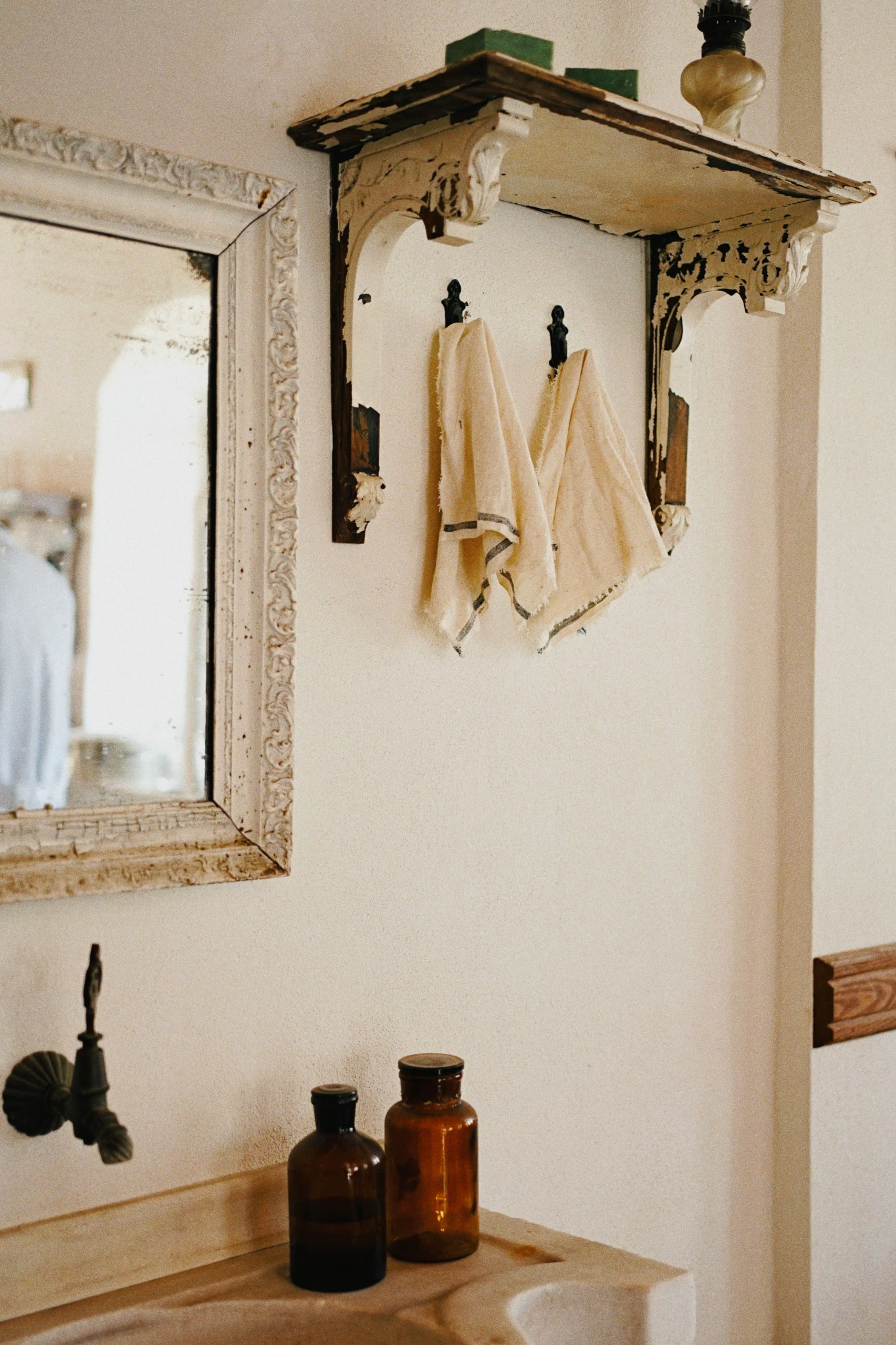 Vintage-style small bathroom with distressed wood accents, featuring on-the-wall hooks holding hand towels beneath an ornate shelf. An inspiring example of space-saving small bathroom ideas.