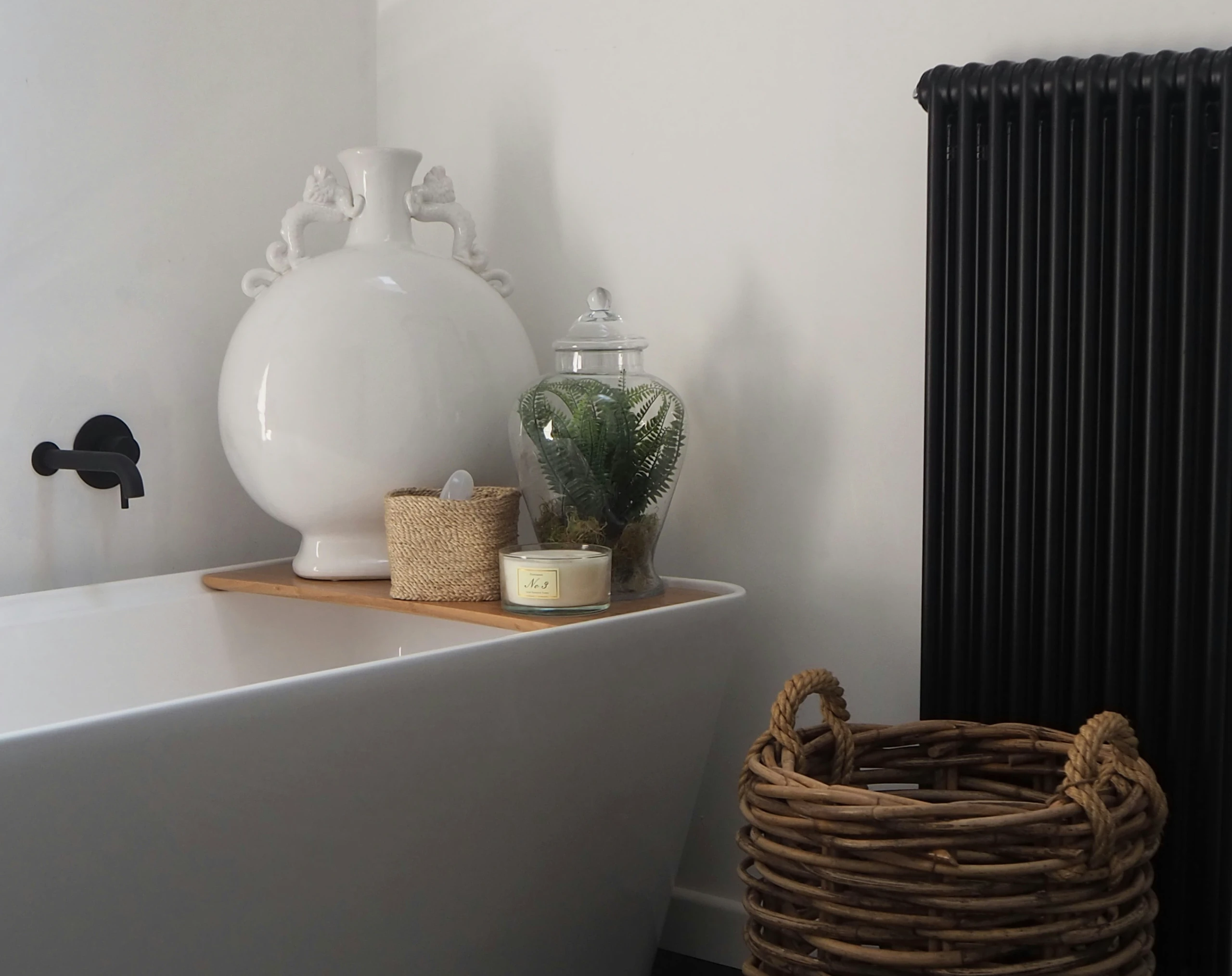 A stylish small bathroom setup featuring a modern white bathtub with a wooden bath tray holding decorative items like a large white vase, a glass jar with greenery, a candle, and a small woven container. To the right, a large wicker basket sits on the floor next to a sleek black radiator—an inspiring example of small bathroom ideas on a budget that incorporates baskets for storage and organization in a functional yet aesthetic way.
