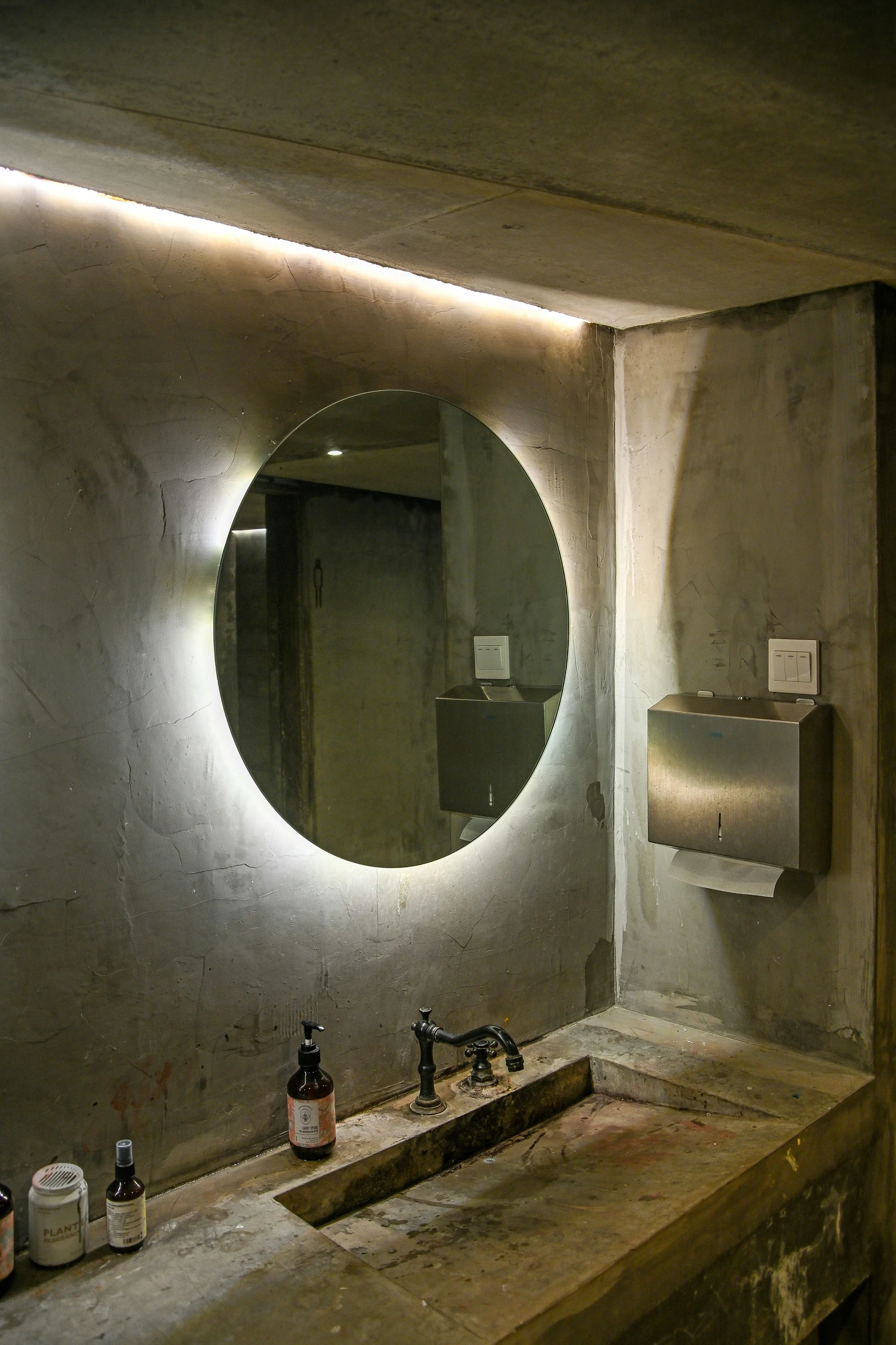 Industrial-style bathroom showcasing small bathroom ideas on a budget, featuring a round mirror with clever lighting from behind to enhance the ambiance and make the space feel larger. The raw concrete textures, minimalist sink, and practical wall-mounted accessories offer a cost-effective yet stylish design solution.