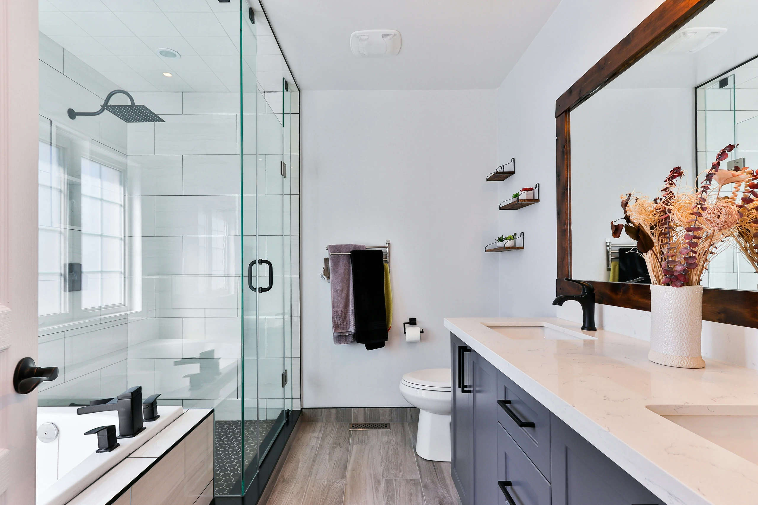 A bright and stylish bathroom showcasing small bathroom ideas on a budget, featuring sleek glass shower doors that help open up the space. The design includes a dual-sink vanity with a marble-look countertop, black fixtures, and a large framed mirror. Light wood-look flooring and floating shelves with small plants add warmth and functionality to this modern, space-efficient layout.