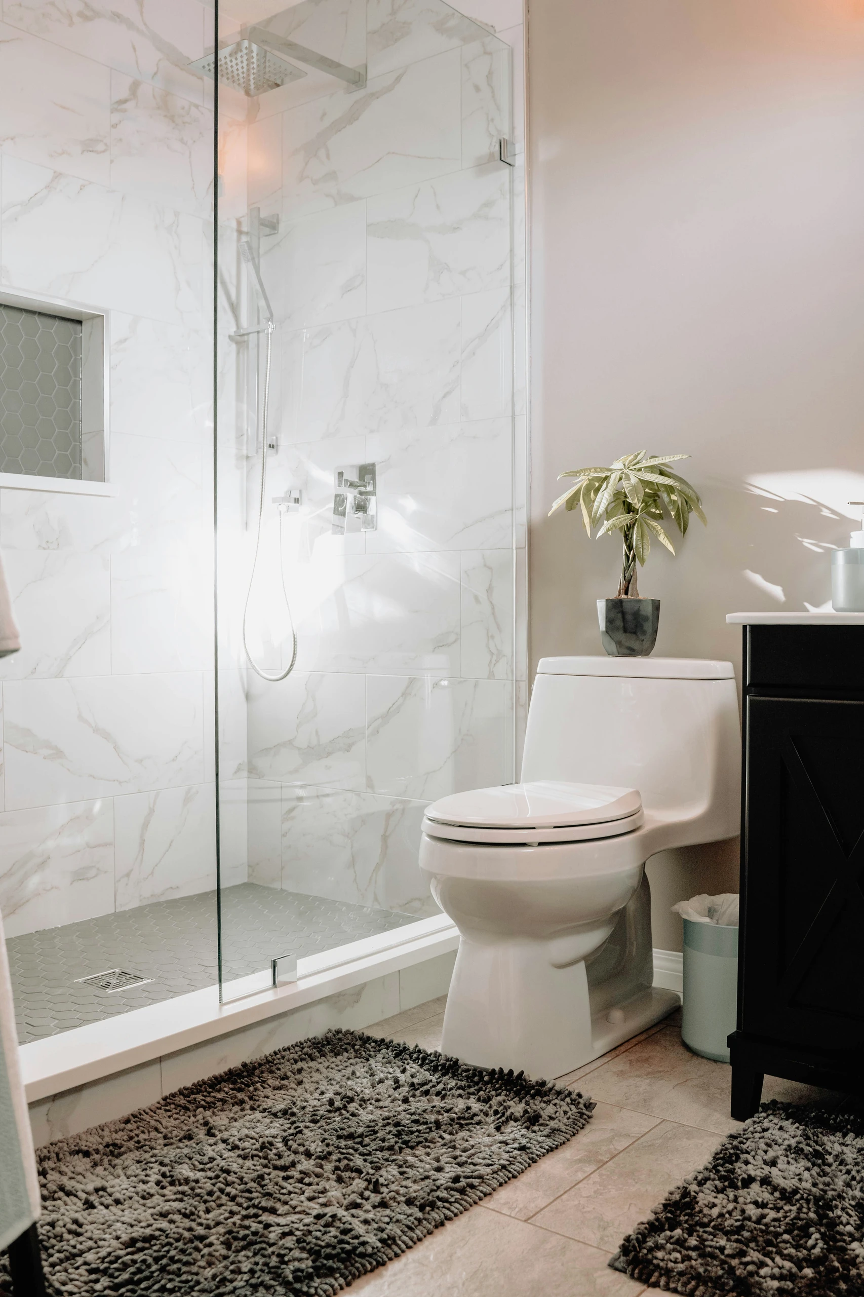 A stylish and functional bathroom showcasing small bathroom ideas on a budget, featuring light colors to enhance the sense of space. The design includes a sleek white toilet, a modern glass-enclosed shower with marble-look tiles, and soft gray textured rugs. A compact vanity and a small potted plant add personality without overwhelming the room, while the use of natural light creates an airy, clean ambiance.