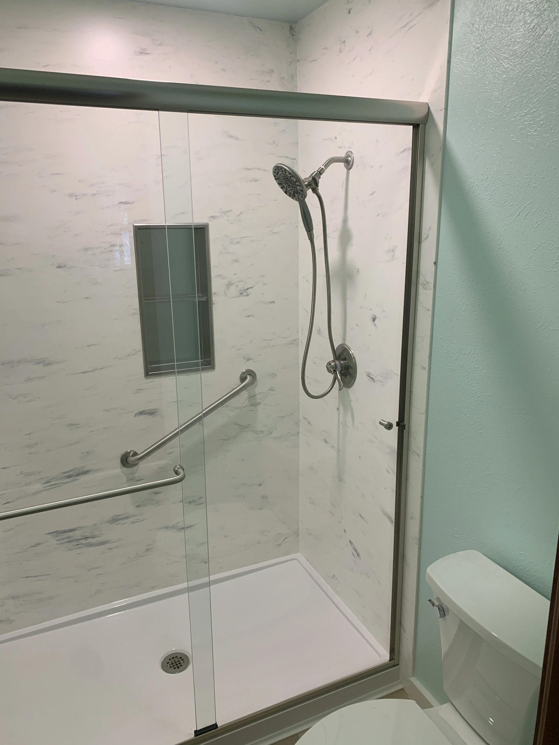 A compact and functional bathroom showcasing small bathroom ideas on a budget, featuring sleek sliding shower doors and a minimalist shower setup with a handheld showerhead and built-in grab bars. The light wall tones and marble-effect panels create a clean, spacious feel perfect for maximizing style in limited spaces.