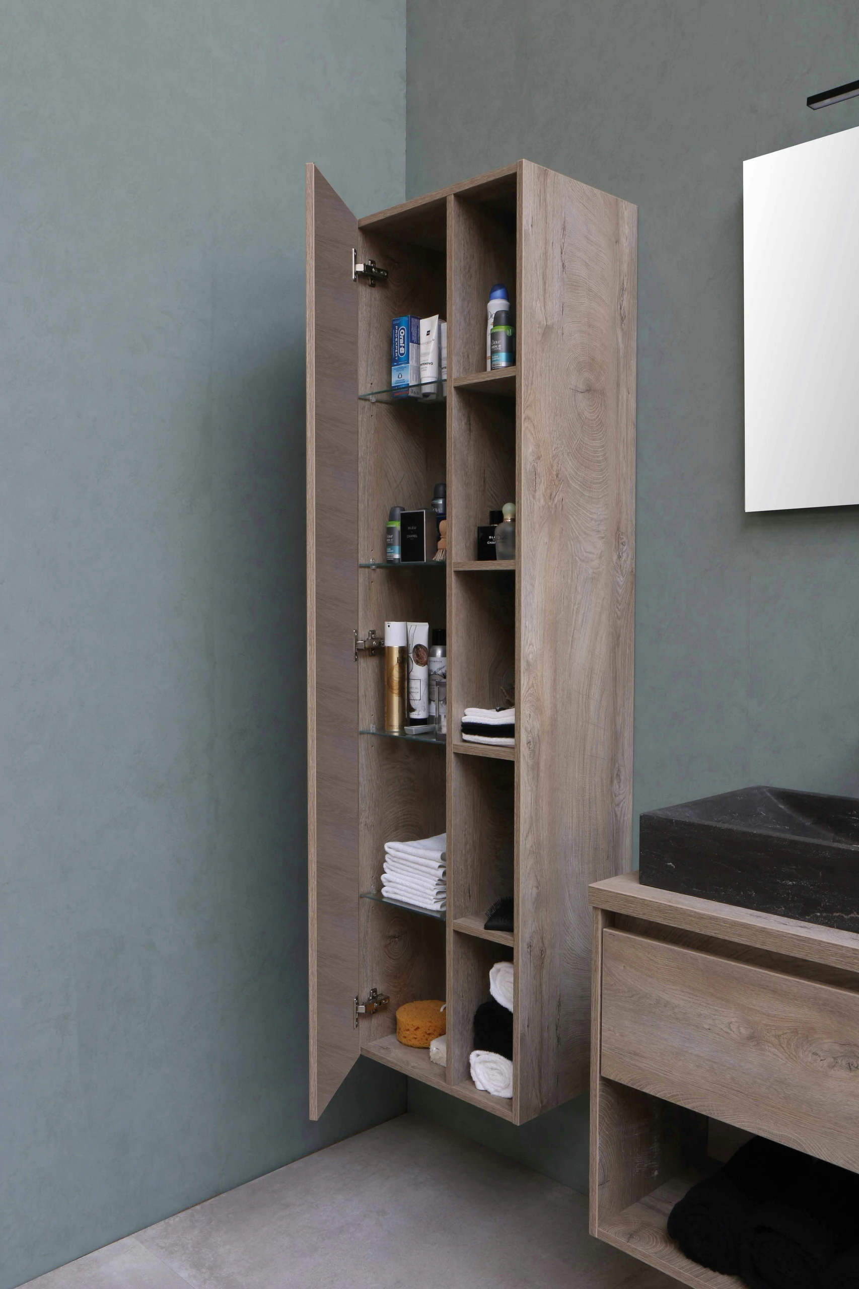 A stylish small bathroom featuring a tall, slim wooden cabinet with open shelves and a door concealing part of the contents. The vertical storage unit holds neatly folded towels, grooming products, and toiletries, maximizing space without overwhelming the room. Adjacent to it is a matching vanity with a dark stone sink and additional open shelving below. This image showcases small bathroom ideas on a budget with smart vertical storage ideas to keep essentials organized while maintaining a clean, modern aesthetic.