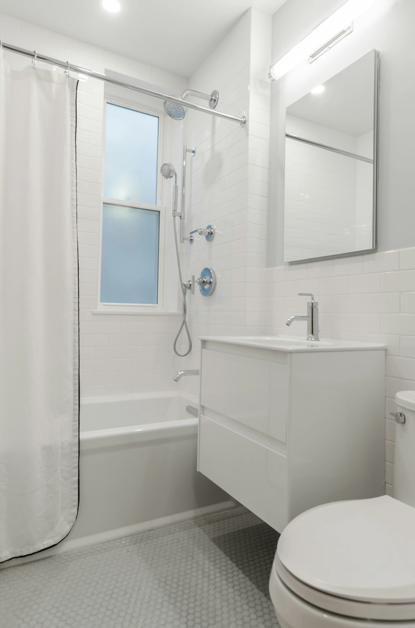 A compact and modern bathroom showcasing small bathroom ideas on a budget, featuring a sleek wall-mounted wash basin that maximizes floor space. The design includes white subway tiles, a combination bathtub and shower with chrome fixtures, and a simple shower curtain. Light gray hexagonal floor tiles and bright overhead lighting enhance the clean, minimalist aesthetic while maintaining functionality in a limited area.