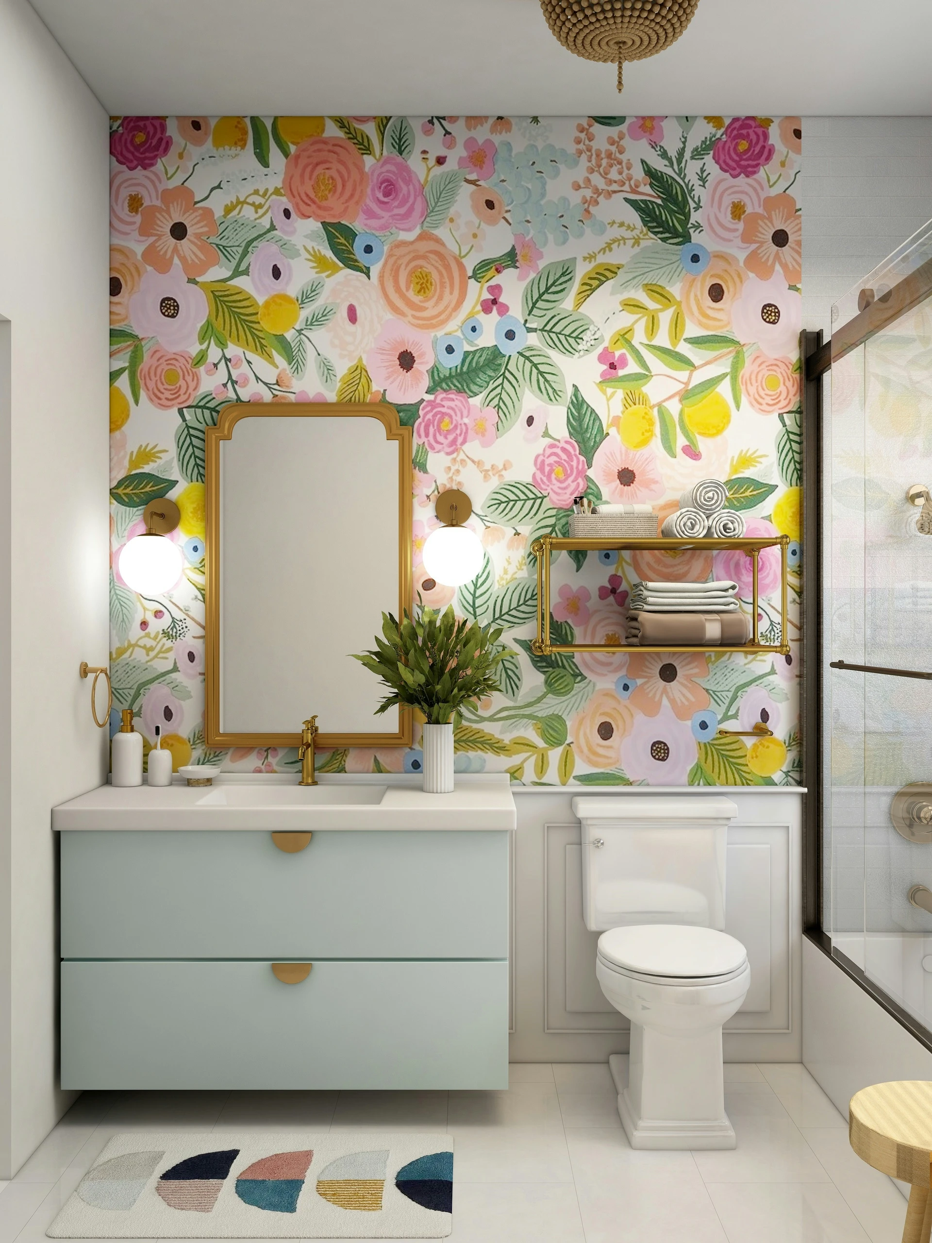 A cheerful and colorful small bathroom featuring a striking floral patterned wallpaper that adds personality and charm to the space. The vanity has soft mint green drawers with gold hardware, paired with a white countertop and gold faucet. Above it, a gold-framed mirror is flanked by modern globe sconces. A shelf above the toilet provides extra towel storage, and a playful rug adds warmth to the floor. This setup showcases small bathroom ideas on a budget, using patterned wallpaper as a cost-effective way to transform and brighten the room.
