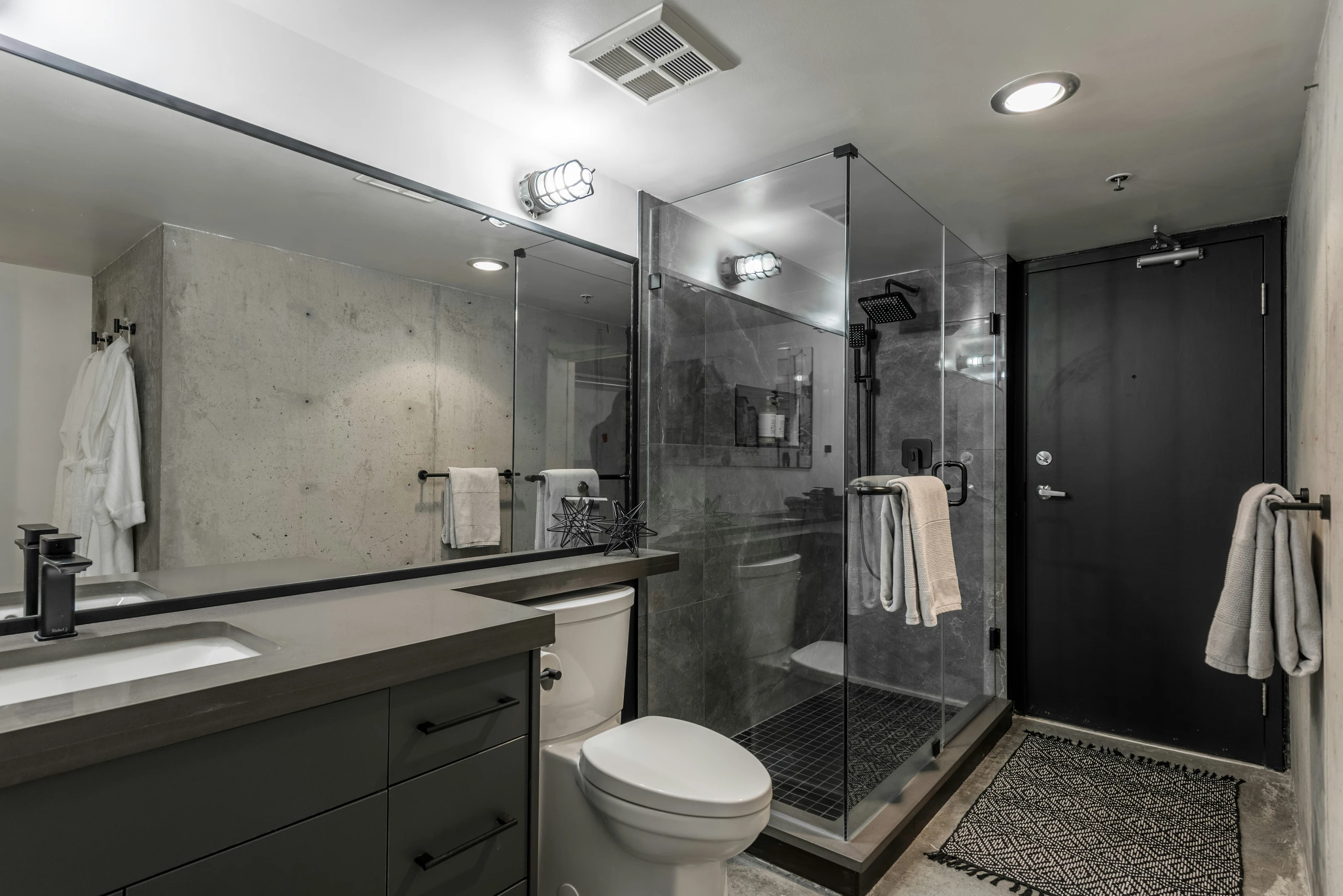 A modern, space-efficient bathroom featuring small bathroom ideas on a budget, including a wide mirror that enhances the sense of space and reflects natural and artificial light. The design pairs a glass-enclosed shower with dark tile, a compact vanity, and minimalist fixtures in black and gray tones, offering a clean, contemporary look suitable for various interior styles.