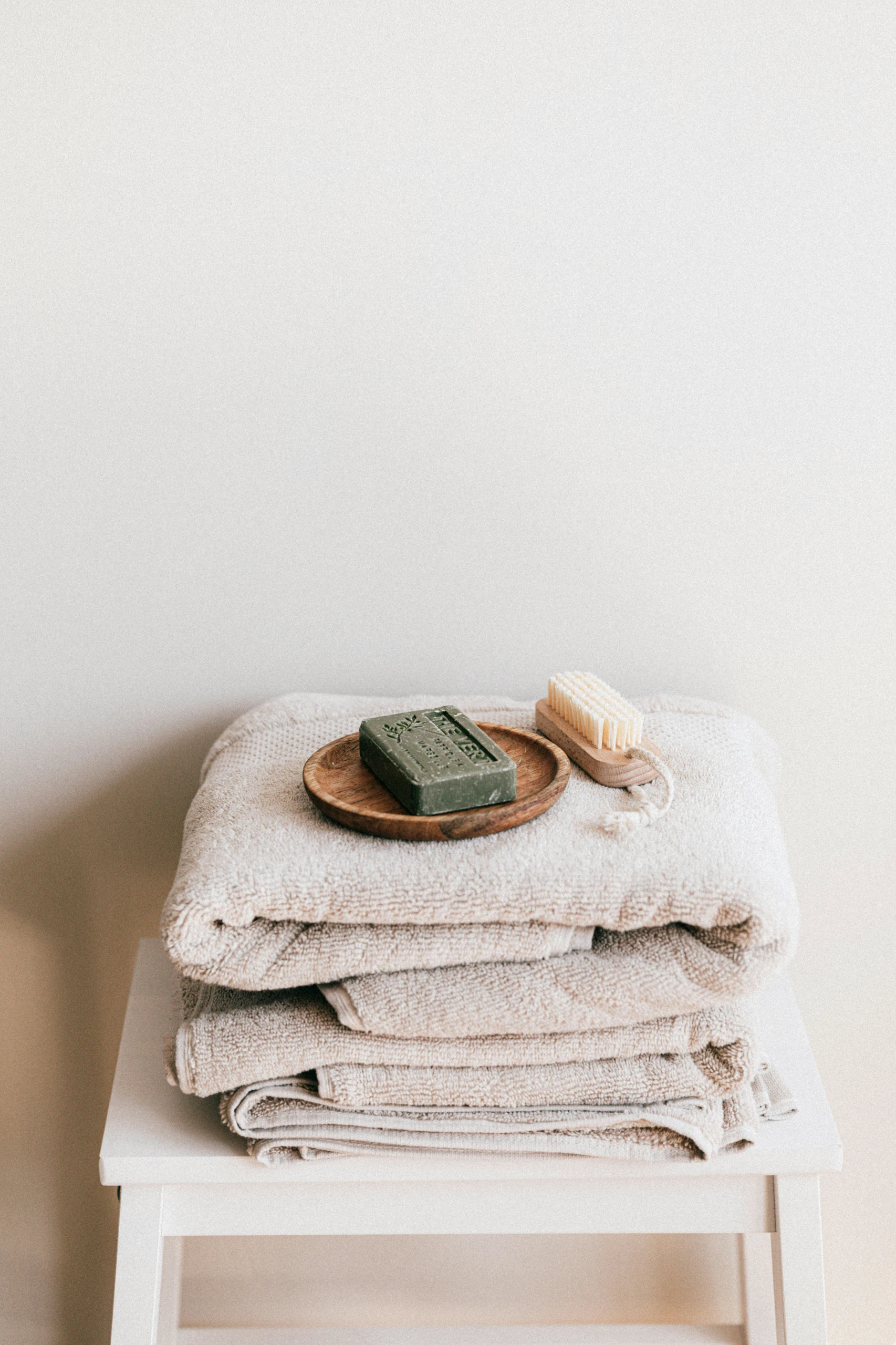 Stack of soft beige towels topped with a natural soap bar and wooden brush on a small white stool, illustrating small bathroom ideas with the use of cozy textiles for a warm, spa-like atmosphere.
