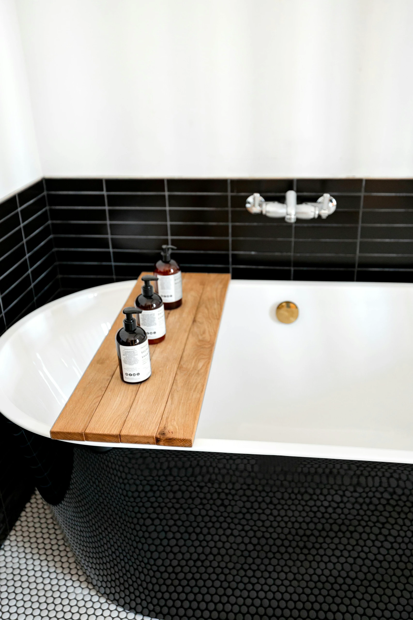Freestanding bathtub in a stylish small bathroom, featuring black penny round special tiles and a wooden bath tray with minimalist soap dispensers. An elegant touch for modern small bathroom ideas.