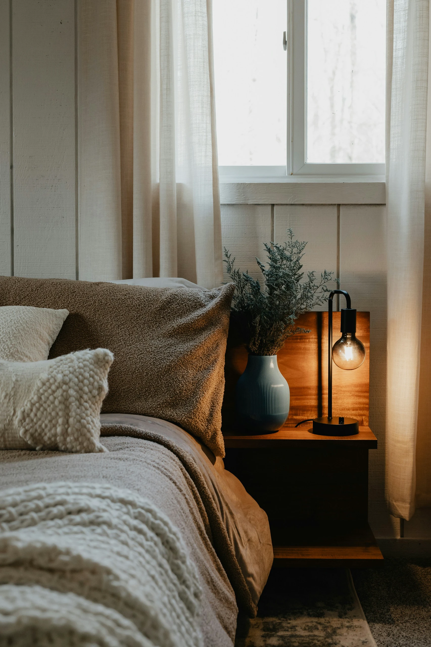 A warm and inviting example of small bedroom ideas with storage, highlighting a floating nightstand made of wood. Positioned next to a cozy bed with textured pillows and blankets, the nightstand features a blue vase with greenery and a sleek modern lamp, demonstrating how floating nightstands can maximize floor space in compact rooms.