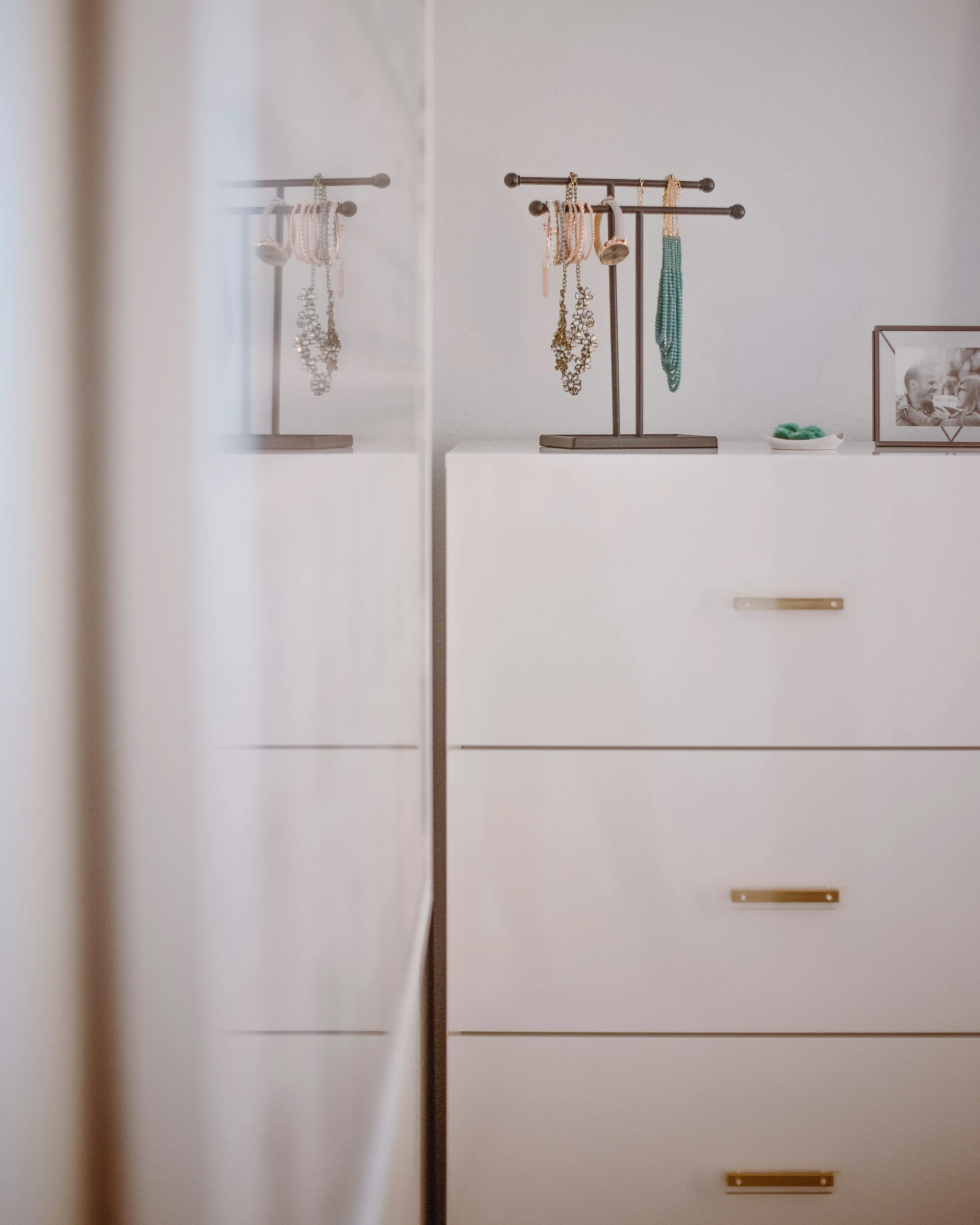 A smart approach to small bedroom ideas with storage, this image features a sleek, white tall dresser with gold handles, used instead of a wider unit to save floor space. Topped with a minimalist jewelry stand and personal items, this vertical storage solution maximizes functionality while maintaining a clean, stylish look.