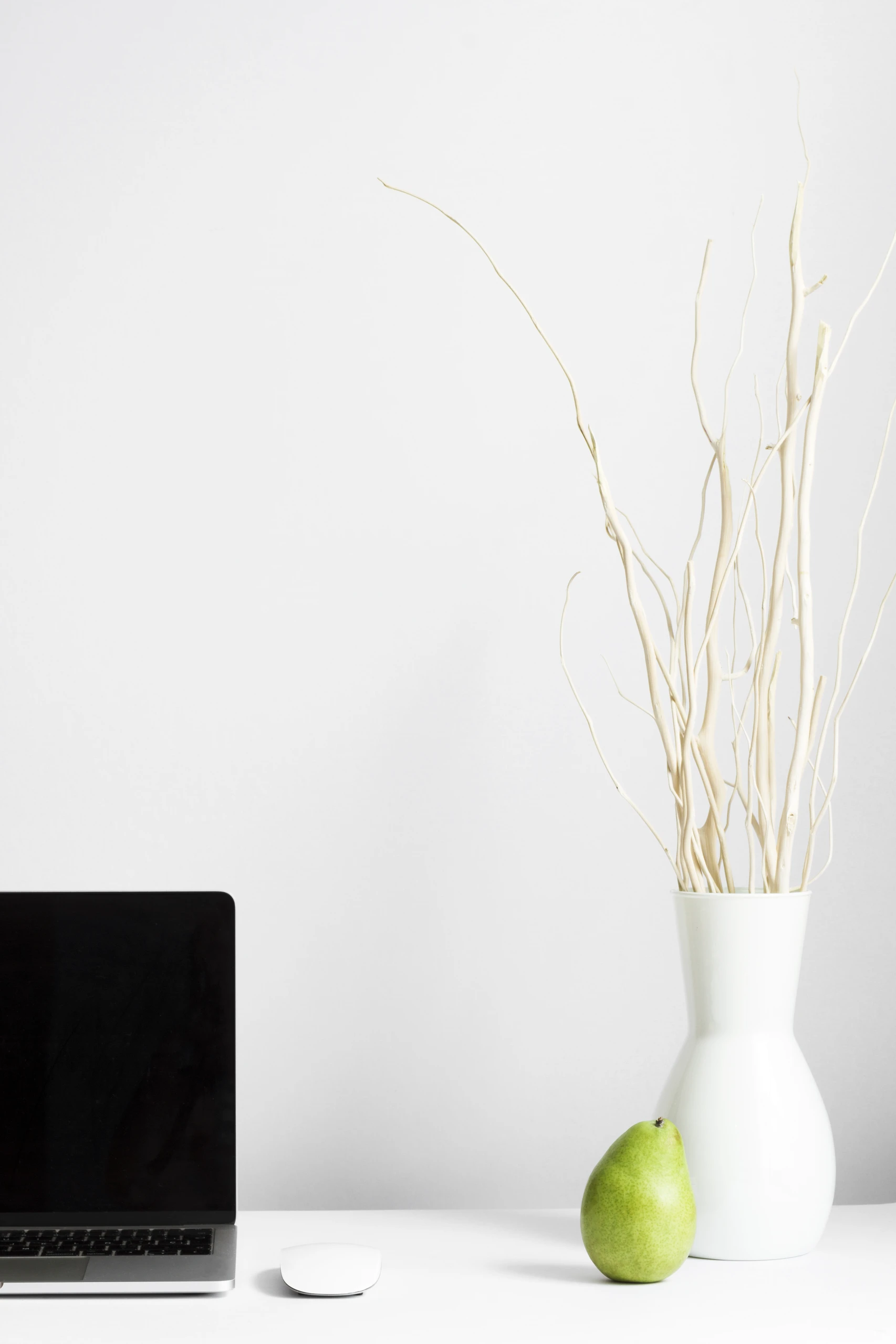 Minimalist workspace in a compact room featuring a fold-down desk with a laptop, wireless mouse, decorative vase with twigs, and a pear. An elegant solution for small bedroom ideas with storage that balances function and aesthetics.