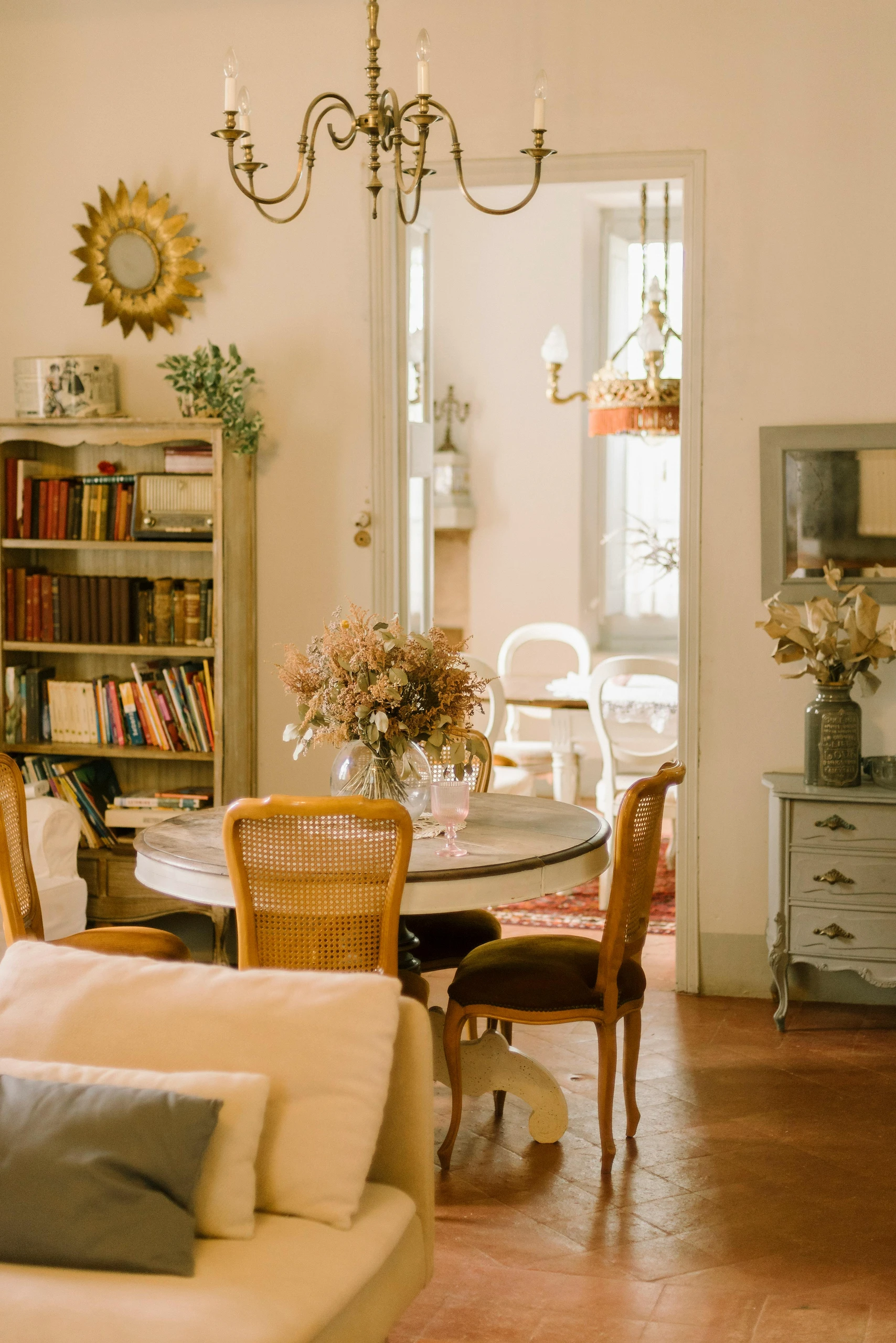 Dining table centerpiece ideas featuring a rustic round table with a glass vase of dried flowers, set beneath an elegant vintage chandelier in a cozy, book-filled room with warm, classic decor.