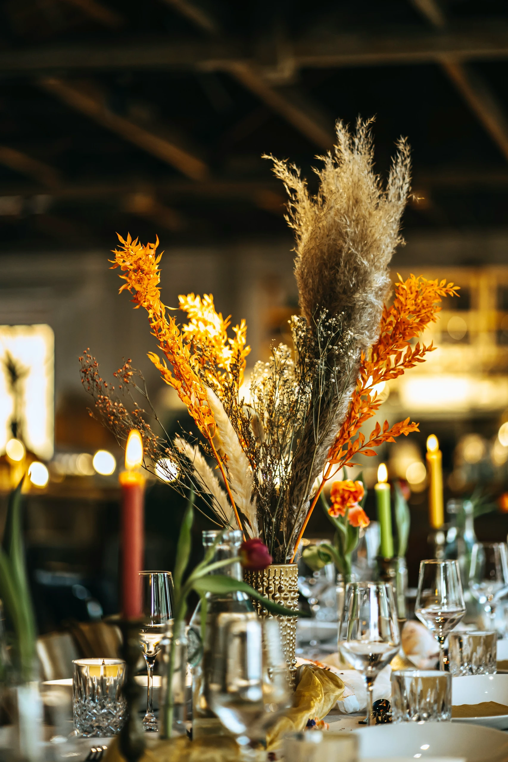 Dining table centerpiece ideas featuring a striking arrangement of dried flowers, including pampas grass and orange foliage, placed in a textured gold vase and surrounded by elegant glassware and glowing candles for a warm, rustic ambiance.