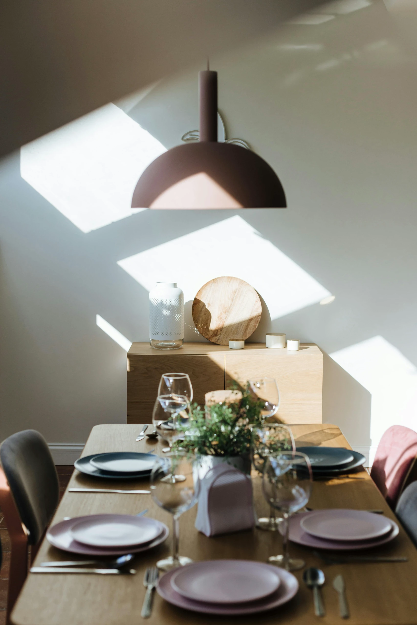 Modern dining table centerpiece ideas featuring a lush green arrangement in the center, complemented by a sleek hanging light above and soft afternoon sunlight casting geometric shadows on the wall.