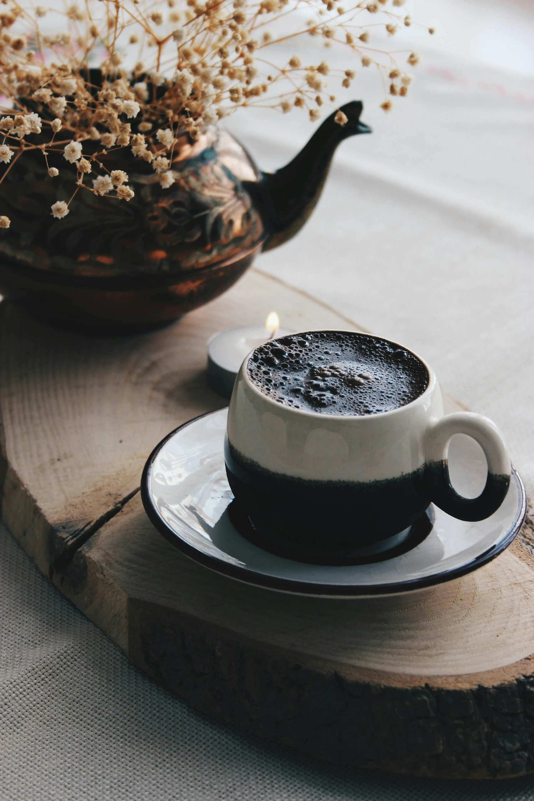 Dining table centerpiece ideas featuring a vintage tea pot filled with delicate dried flowers, placed on a rustic wooden slab alongside a cup of coffee and a small candle for a cozy, inviting atmosphere.