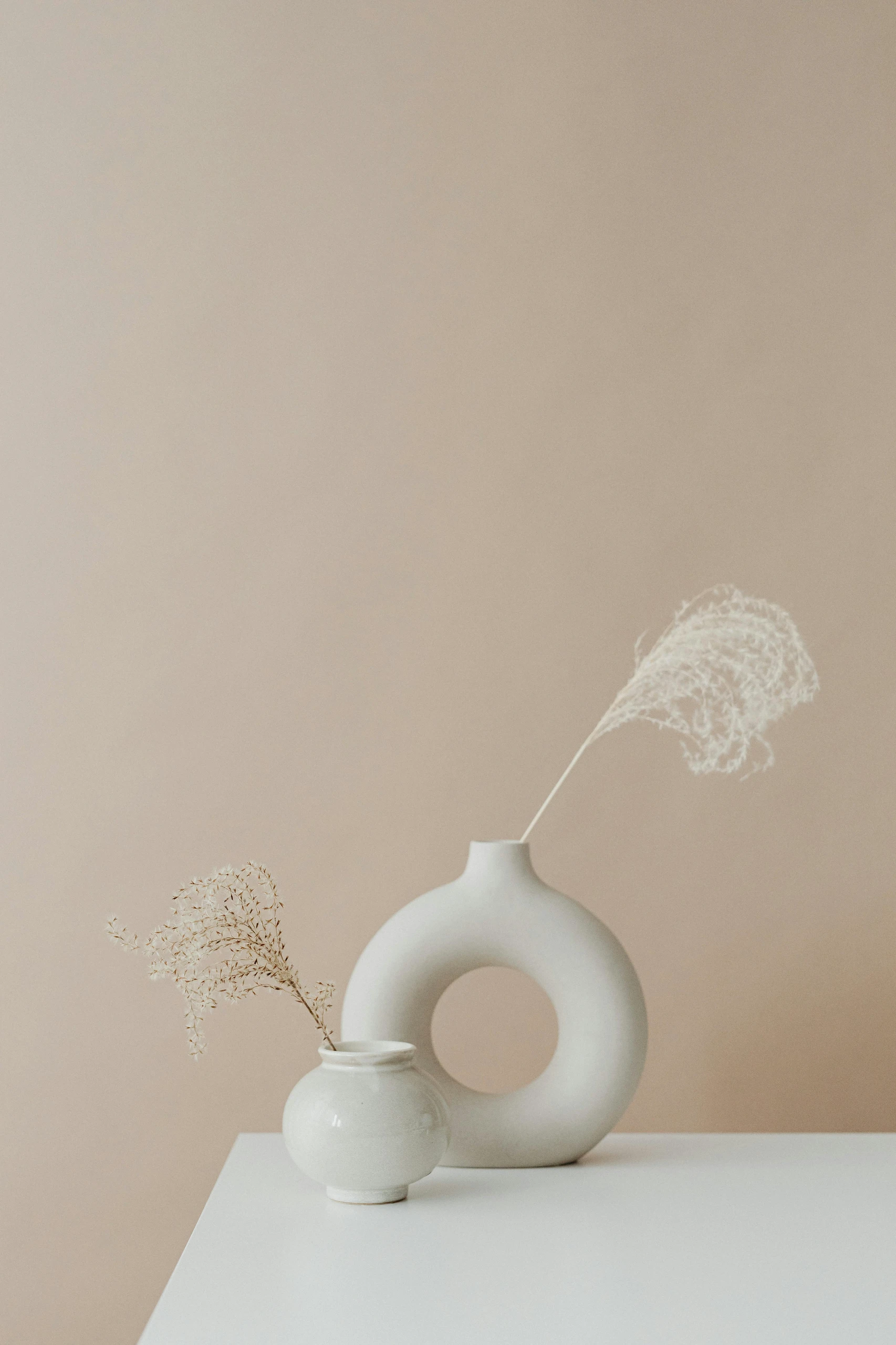 Minimalist dining table centerpiece ideas featuring a unique vase with a hole and a smaller round vase, each holding delicate dried foliage for an elegant, modern look.