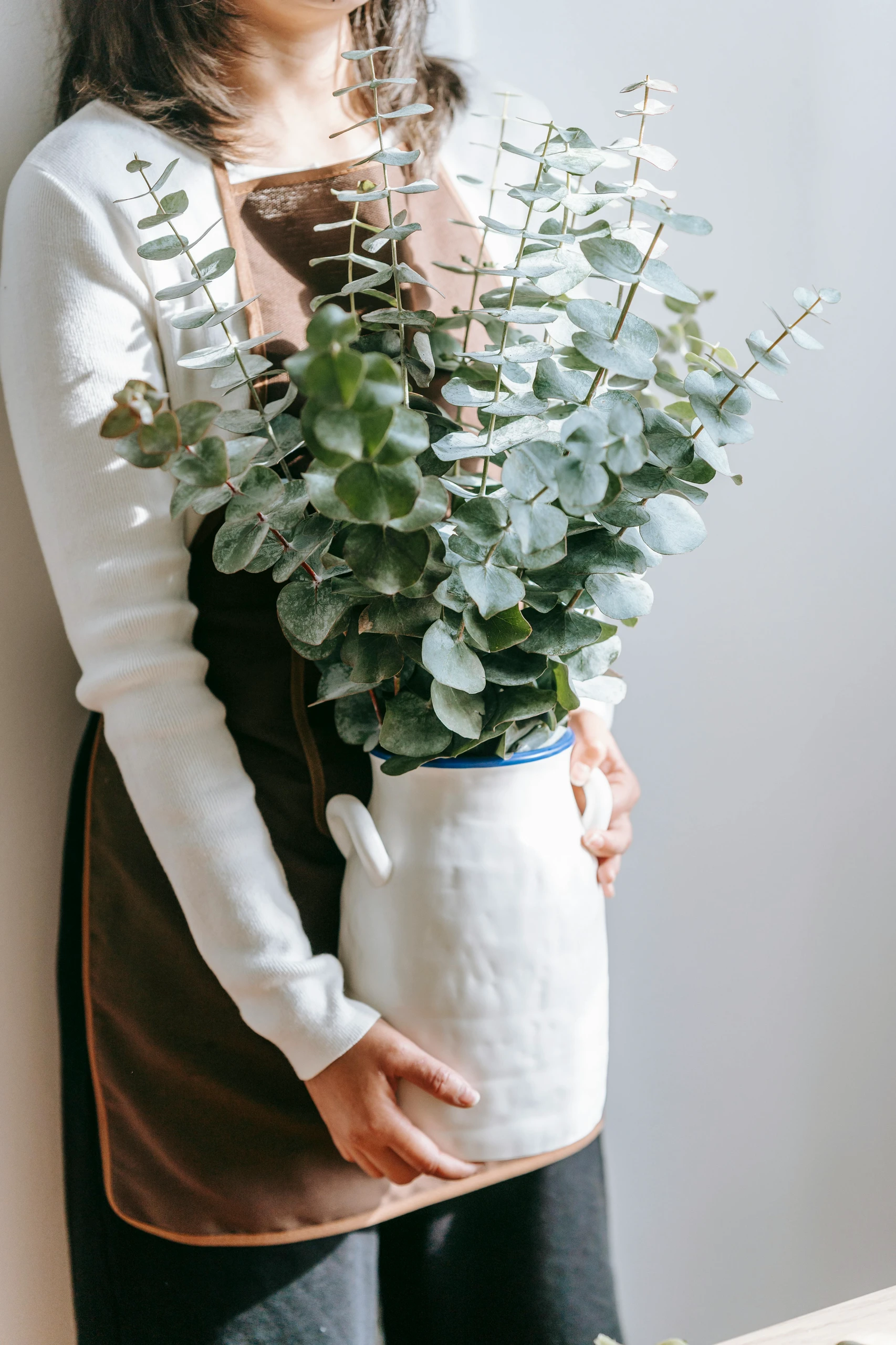Dining table centerpiece ideas featuring fresh greenery in a white ceramic vase, held by a woman in a brown apron, perfect for a minimalist and natural decor style.