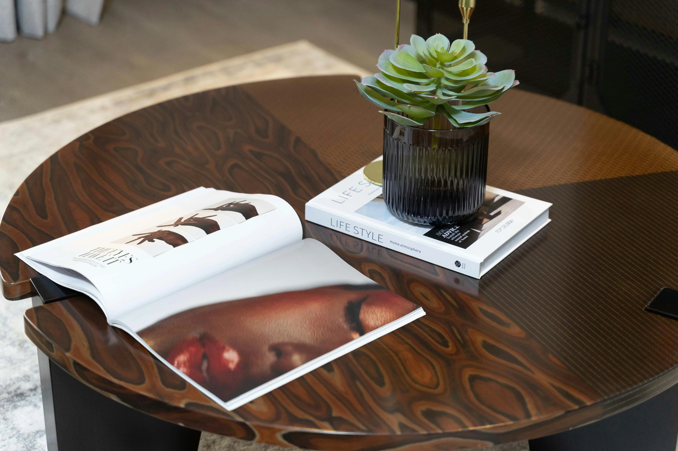 A stylish living room setup demonstrating how to decorate a round coffee table with books and a succulent for a modern, versatile look. The dark wood grain table features an open magazine, stacked lifestyle books, and a textured black pot with a green succulent, blending elegance and simplicity.