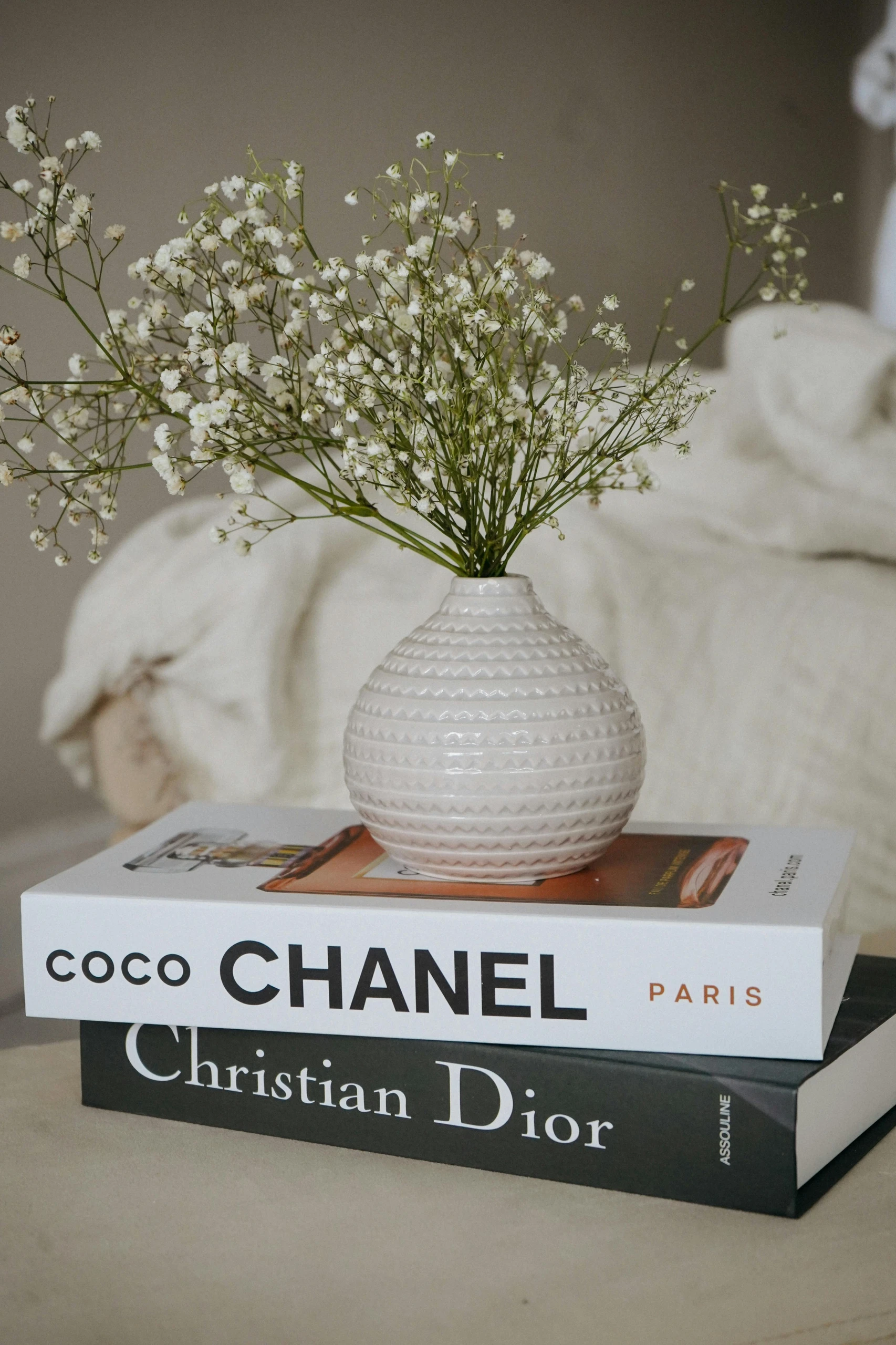 Elegant styling inspiration for how to decorate a round coffee table, featuring a small textured white vase with baby's breath flowers placed atop two stacked fashion books, “Coco Chanel” and “Christian Dior”, for a timeless, minimalist look using books with a small vase on top.