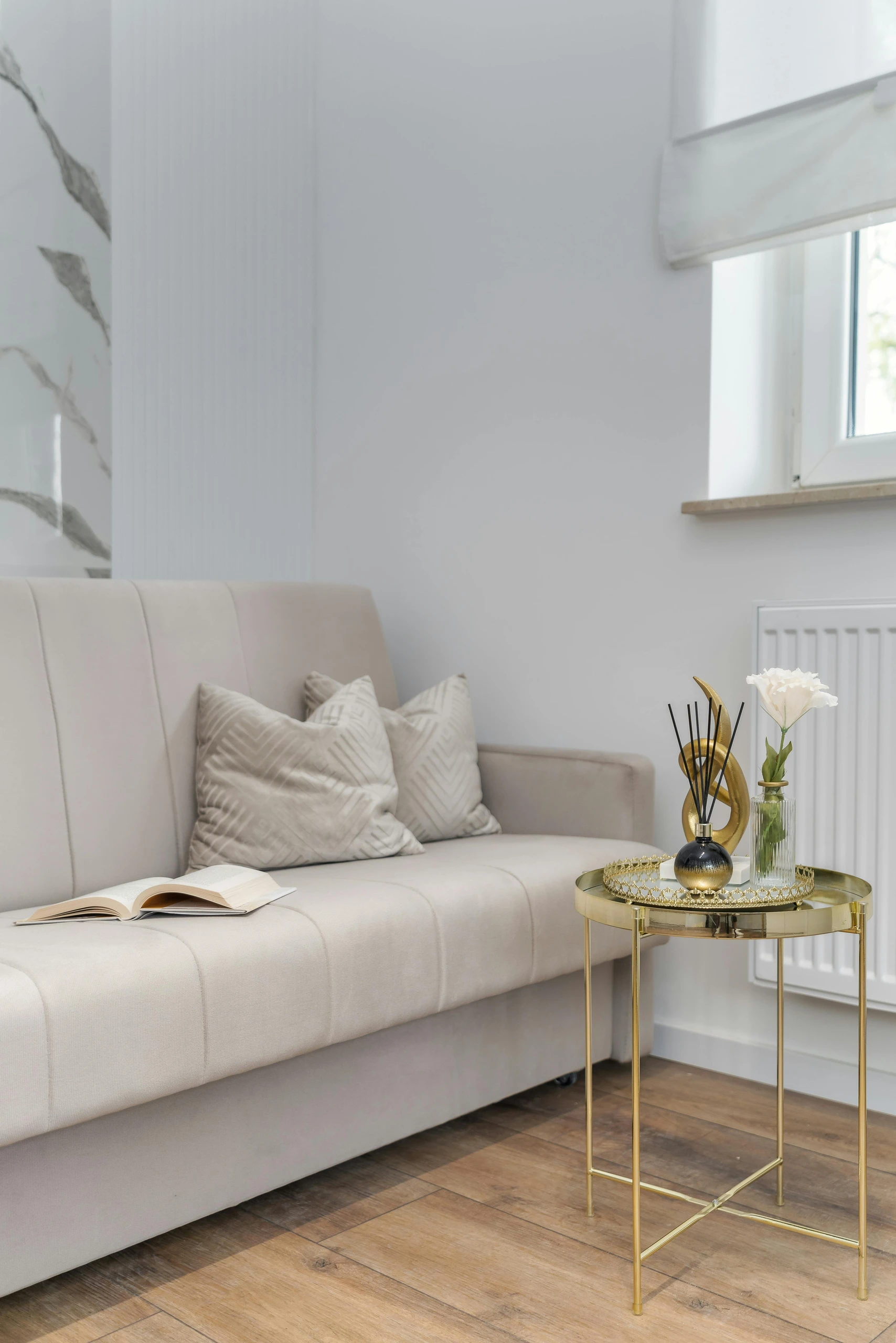 Elegant living room corner showcasing how to decorate a round coffee table with gold decor pieces for a chic and versatile look. The small metallic table features a gold tray, a sculptural accent, reed diffuser, and a single flower in a glass vase, demonstrating a stylish yet minimalist approach to table styling.