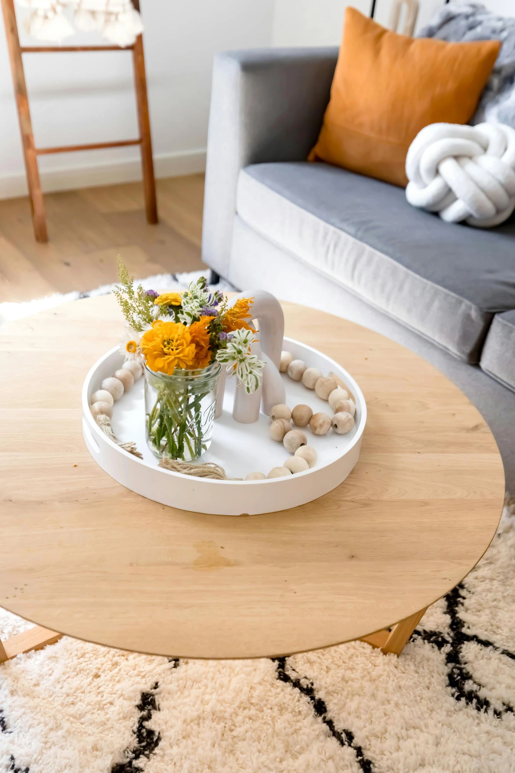 A bright and cozy example of how to decorate a round coffee table using a white tray with flowers and a wood element. The arrangement adds charm and versatility to any living room, combining natural textures with a pop of color for a warm, inviting look.