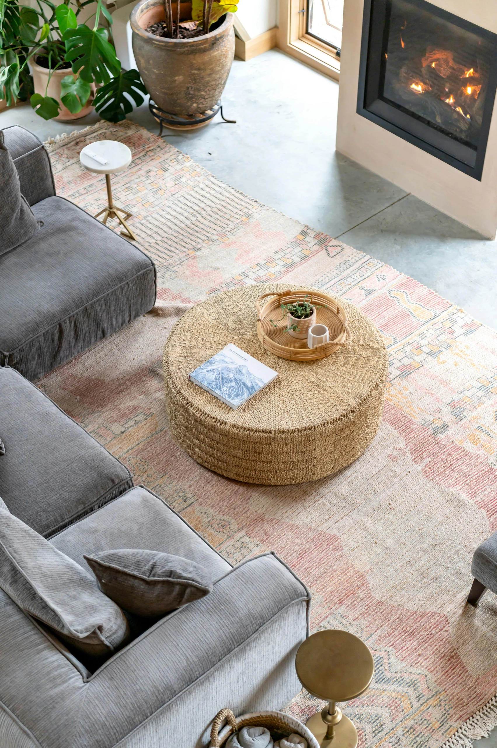 Cozy and versatile take on how to decorate a round coffee table using a woven design and a tray with small plant pot, ceramic mug, and book-perfectly complementing the earthy tones, fireplace warmth, and soft textured rug in a relaxed living space.