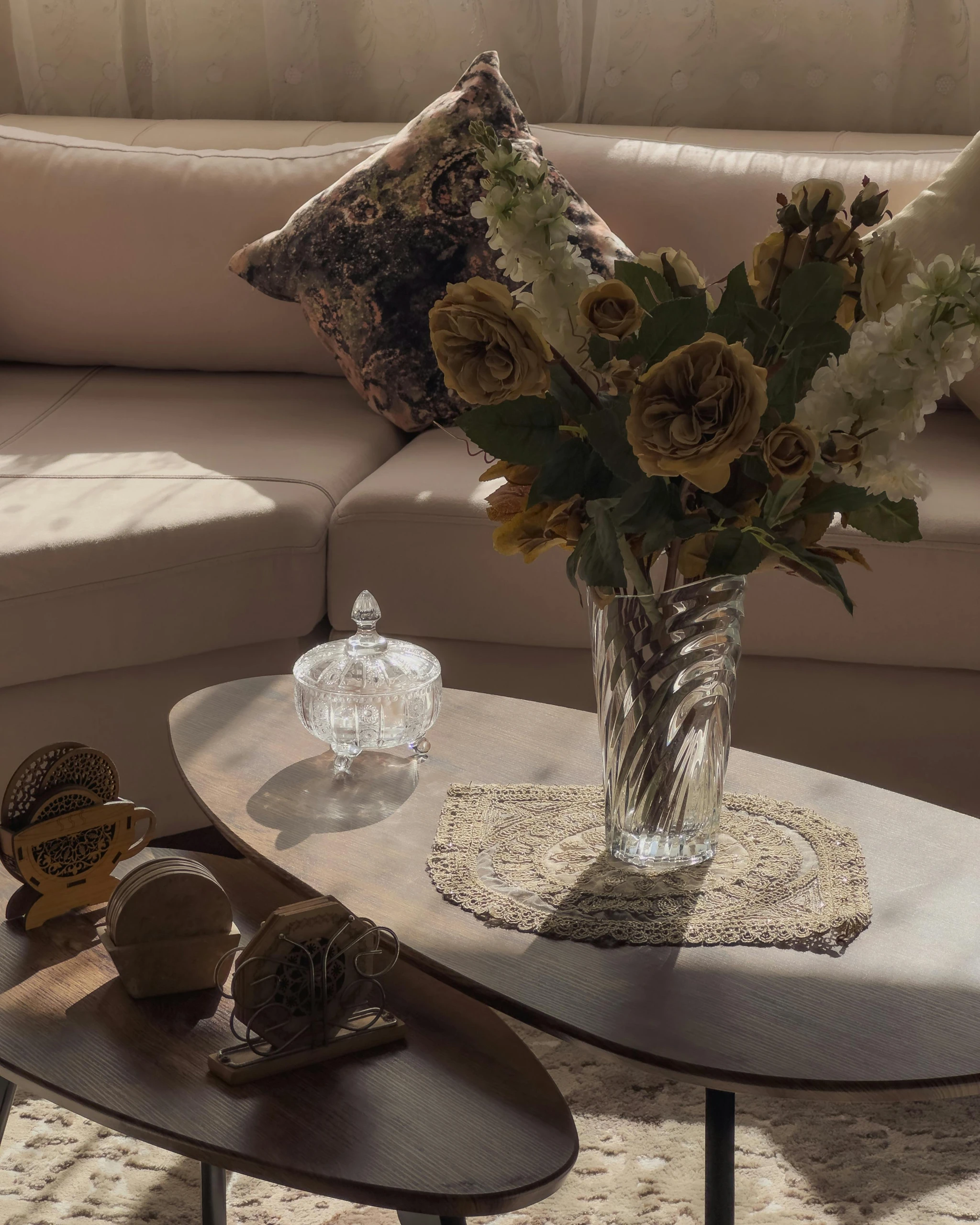 A cozy living room corner featuring a two-pieces-coffee-table with large flower vase and decor elements like a lace doily, a crystal jar, and vintage-style coasters. The warm tones and layered accessories offer inspiration for how to decorate a round coffee table in both classic and modern interiors.