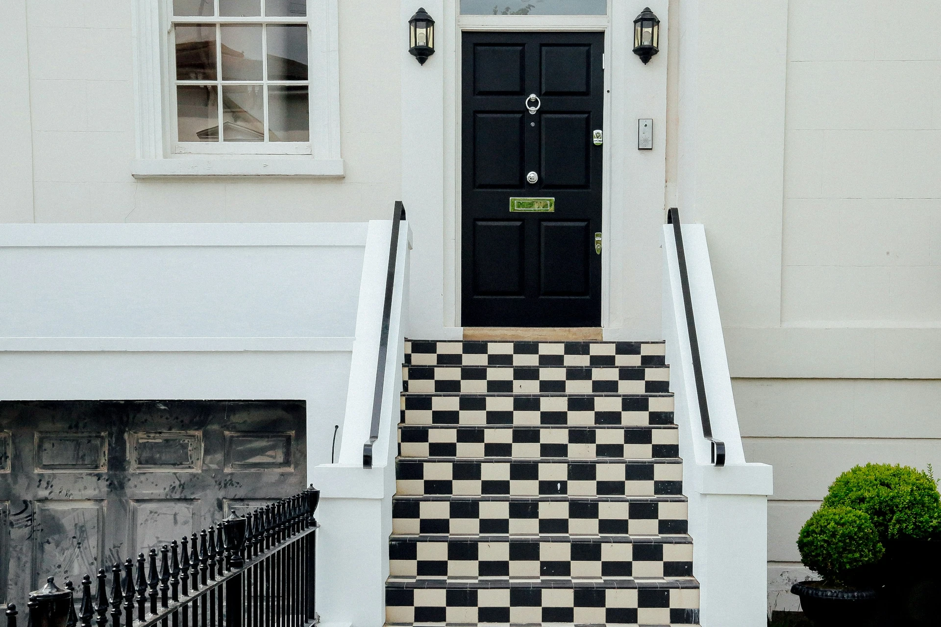 A bold black front door framed by a clean white exterior, with lantern-style lights and a striking checkerboard patterned staircase leading up to the entrance. Perfect for unique ideas for front door decor that incorporate a checkerboard patterned staircase.