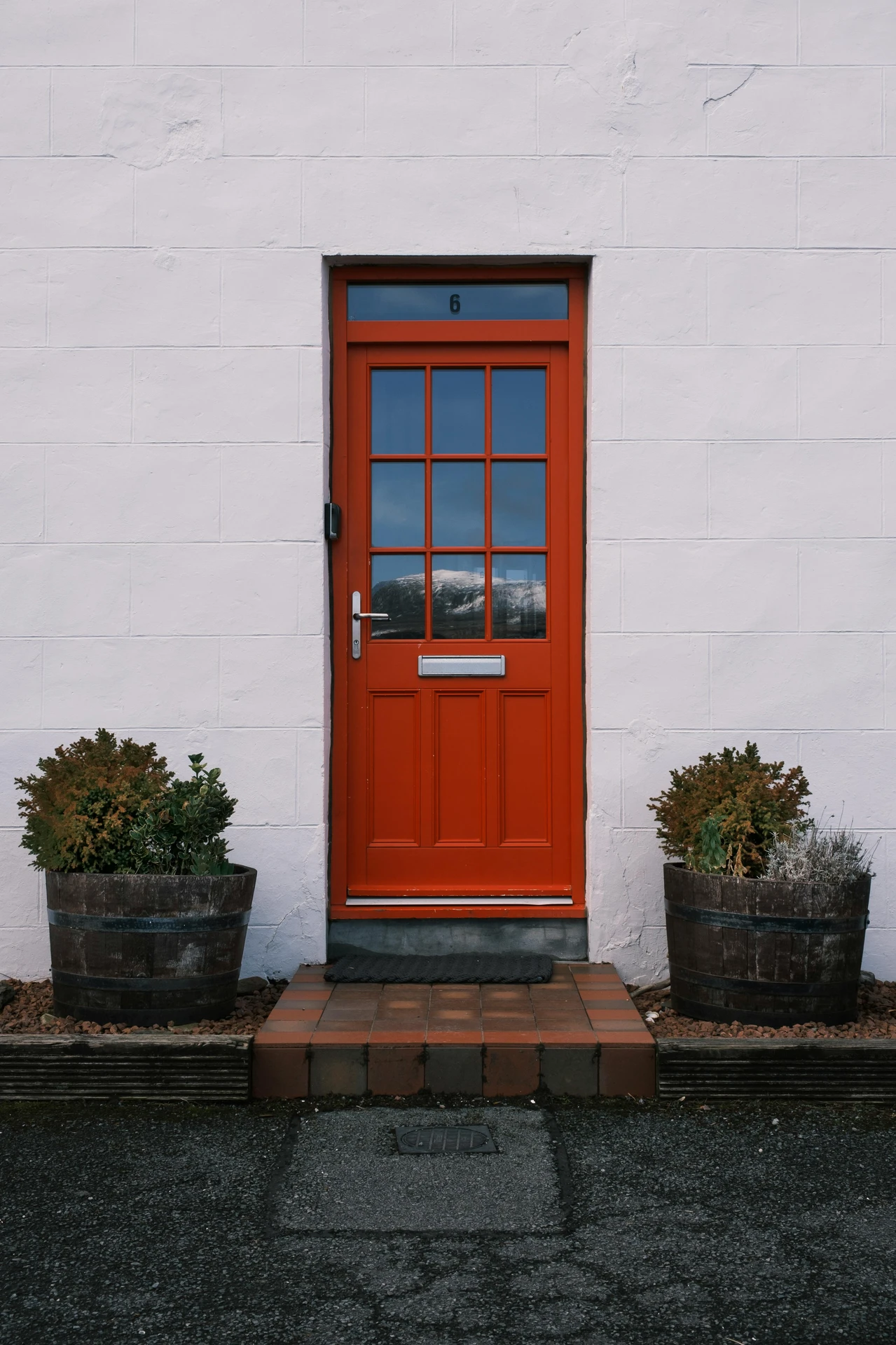 Bold red front door with grid glass panel framed by white exterior walls, flanked by rustic round plant pots. An eye-catching inspiration of ideas for front door decor using color accent & round plant pots for a modern yet approachable style.
