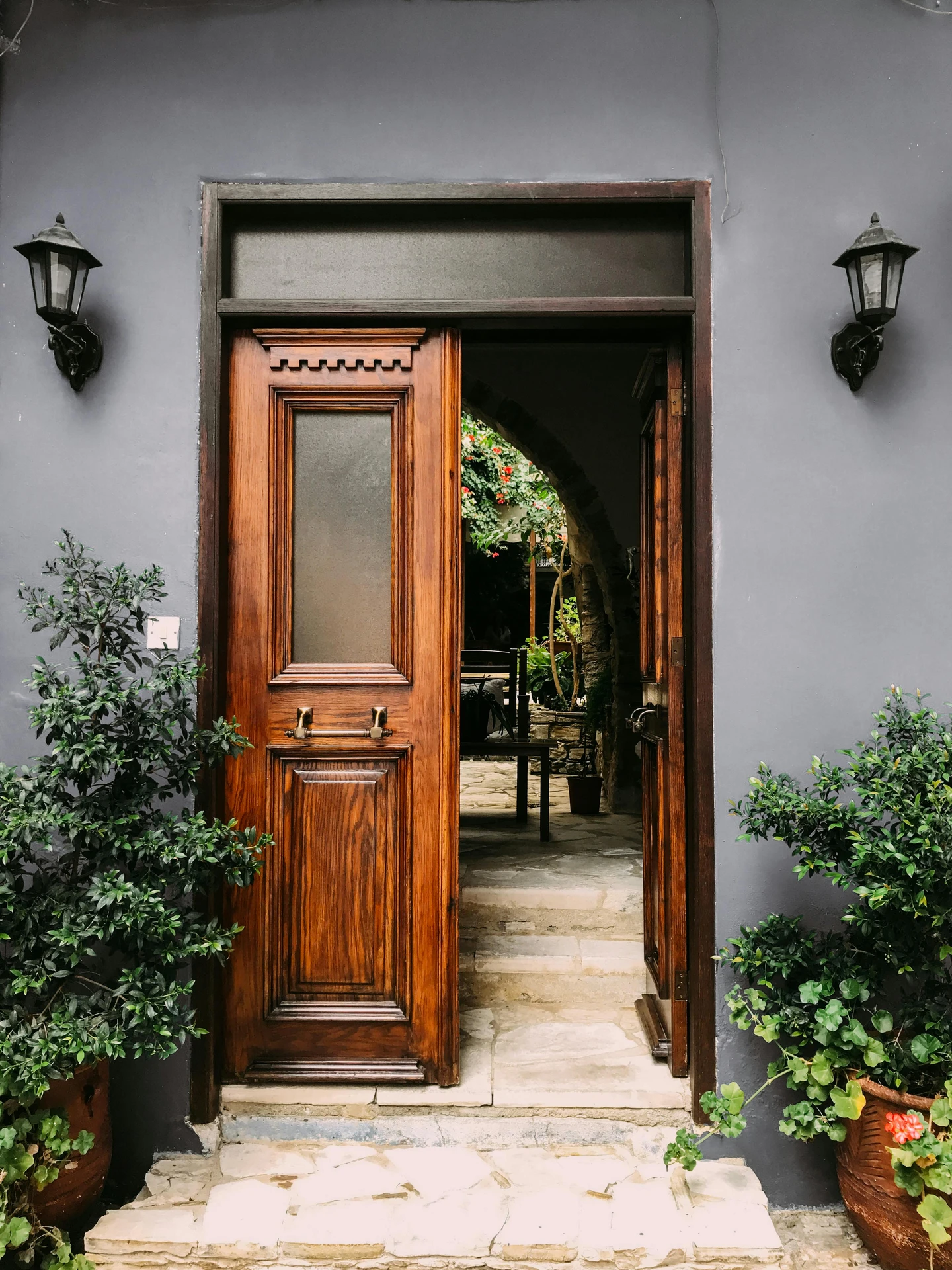 Charming entryway featuring a rustic door made of richly grained wood, framed by matte gray walls and two black wall lanterns, with lush potted greenery on either side. Perfect inspiration for ideas for front door decor with a cozy, rustic door aesthetic.