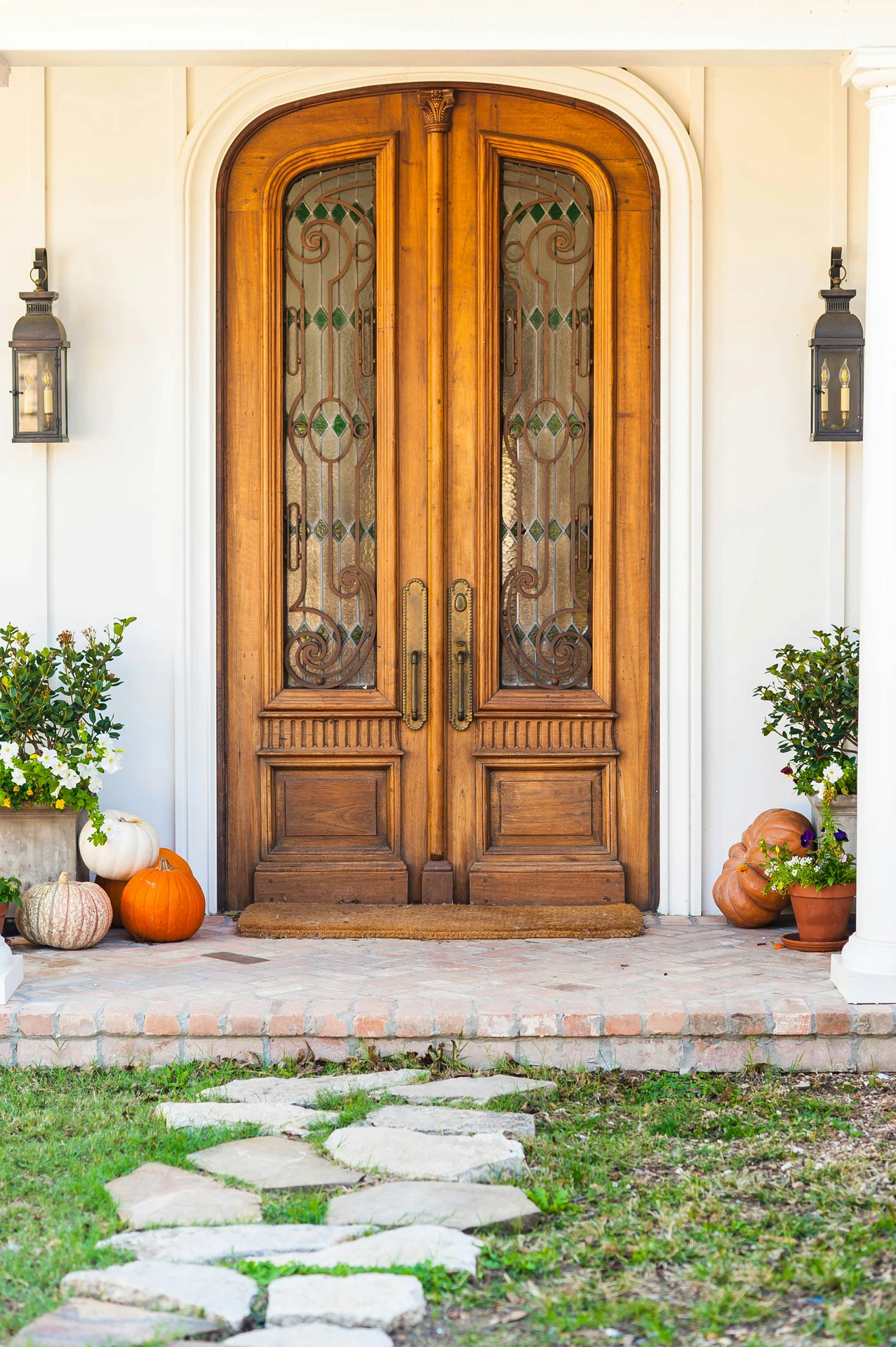 Elegant wooden double doors with ornate glass panels, flanked by potted flowers and a variety of pumpkins in orange, white, and green tones. Nice inspiration for front door decor that highlight cozy and inviting seasonal decor for fall.