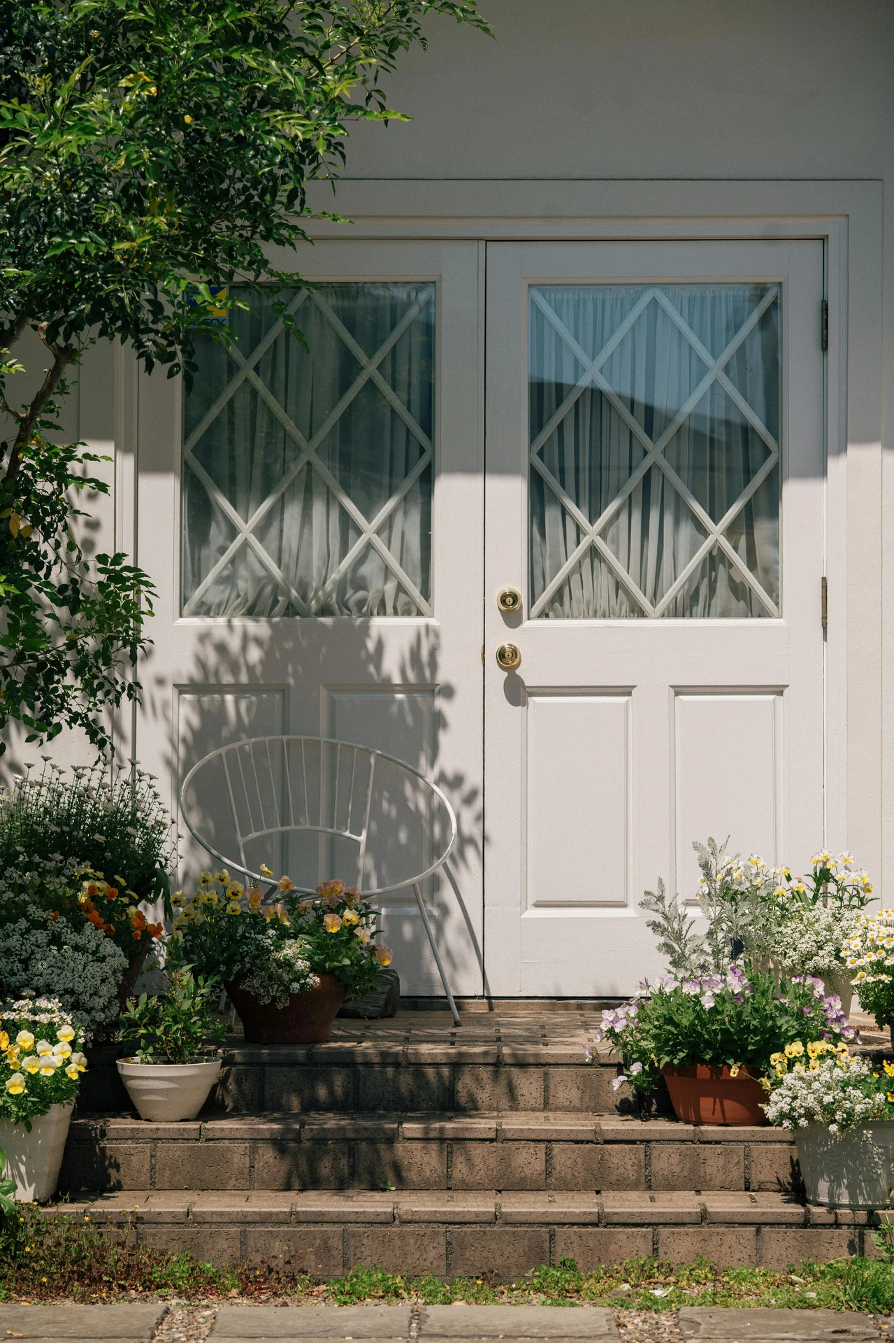 Bright and welcoming front door with white double doors and diamond-patterned glass, framed by lush potted flowers and a simple outdoor seat, ideal for relaxing in the sun. A cozy and versatile inspiration for ideas for front door decor that blends charm, comfort, and natural beauty.