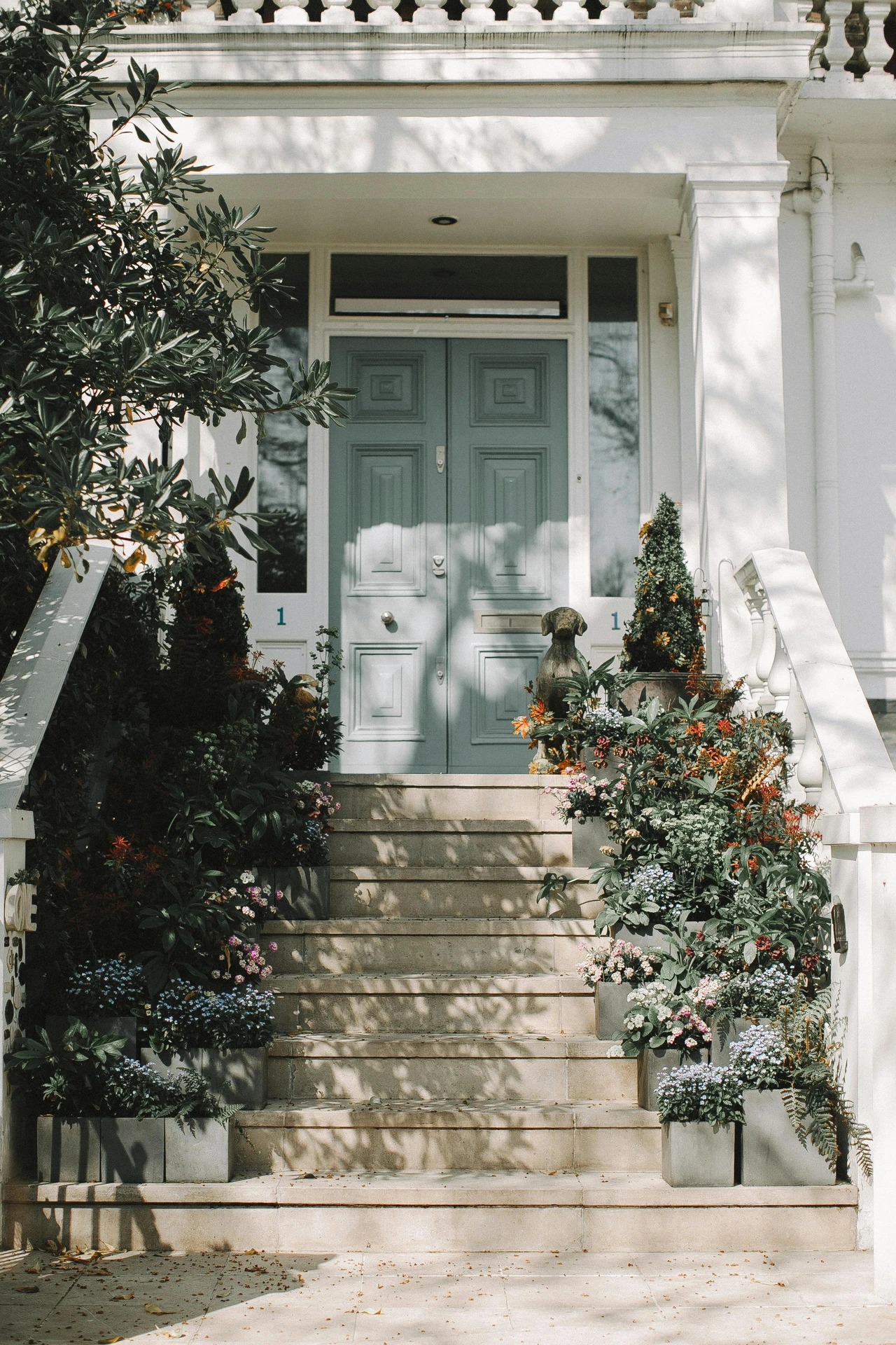A charming front entry featuring square flower boxes on steps, brimming with colorful blooms and greenery, leading to a classic double door in muted teal. This image offers elegant and inviting ideas for front door decor, blending symmetry, seasonal florals, and natural elements for a warm welcome.
