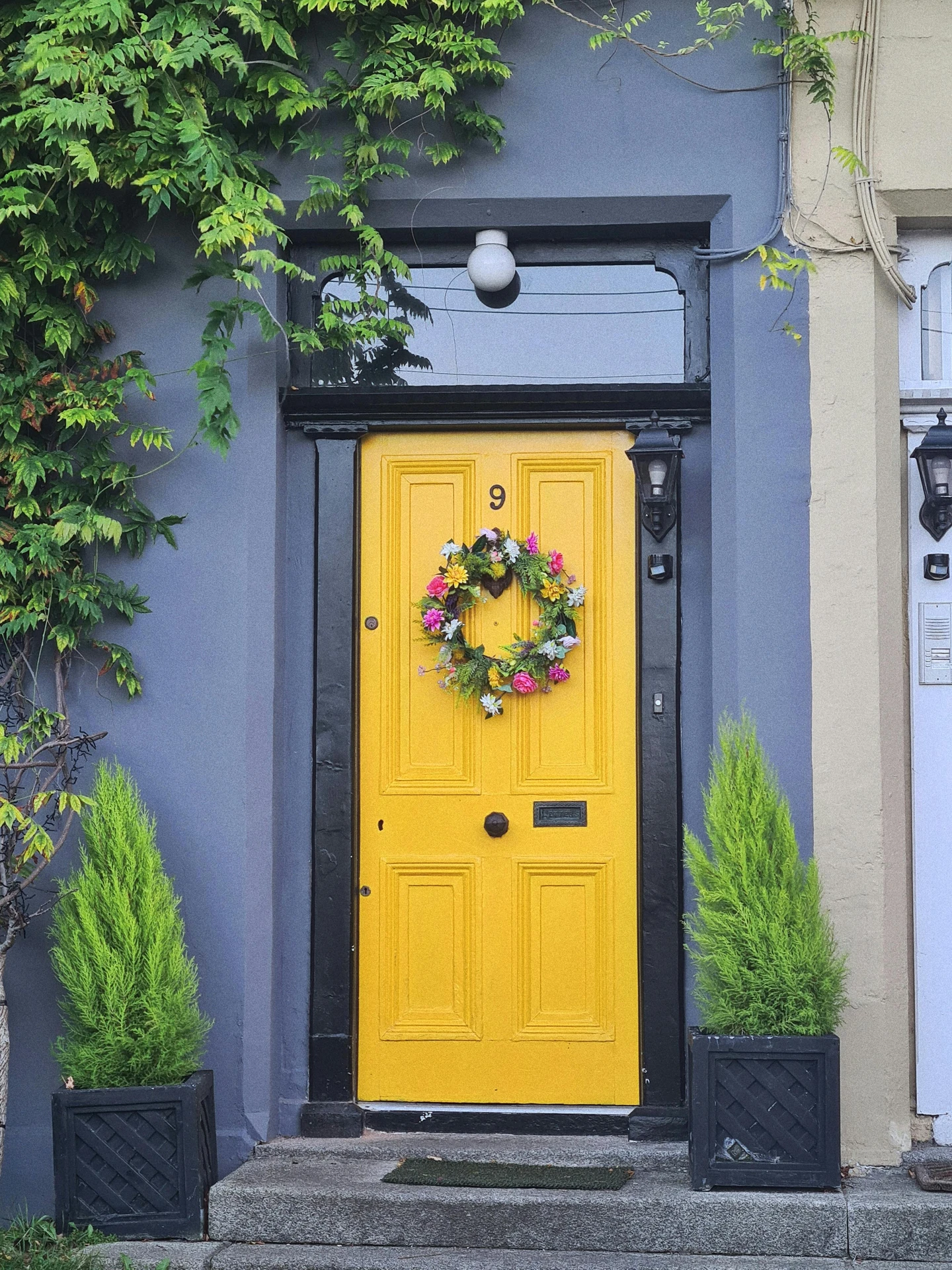 Bright yellow front door framed by a dark gray exterior, decorated with a colorful floral wreath and flanked by two symmetrical potted evergreens, offering vibrant and cheerful ideas for front door decor with a seasonal wreath as the focal point.