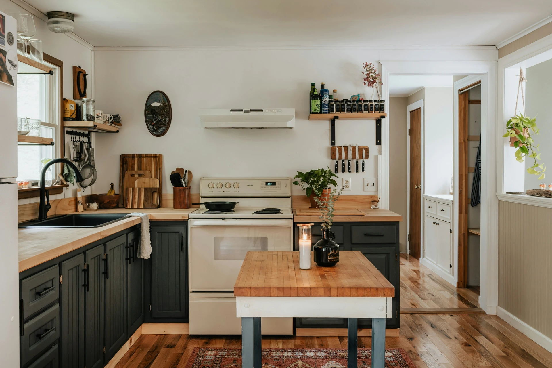 Cozy and rustic kitchen with wood countertops, dark lower cabinets, and a small central island that doubles as a workspace. The island features simple kitchen island decor ideas with a candle and greenery in dark glass vases, adding warmth and charm to the space. Open shelving, hanging utensils, and natural textures complete the inviting, lived-in look.
