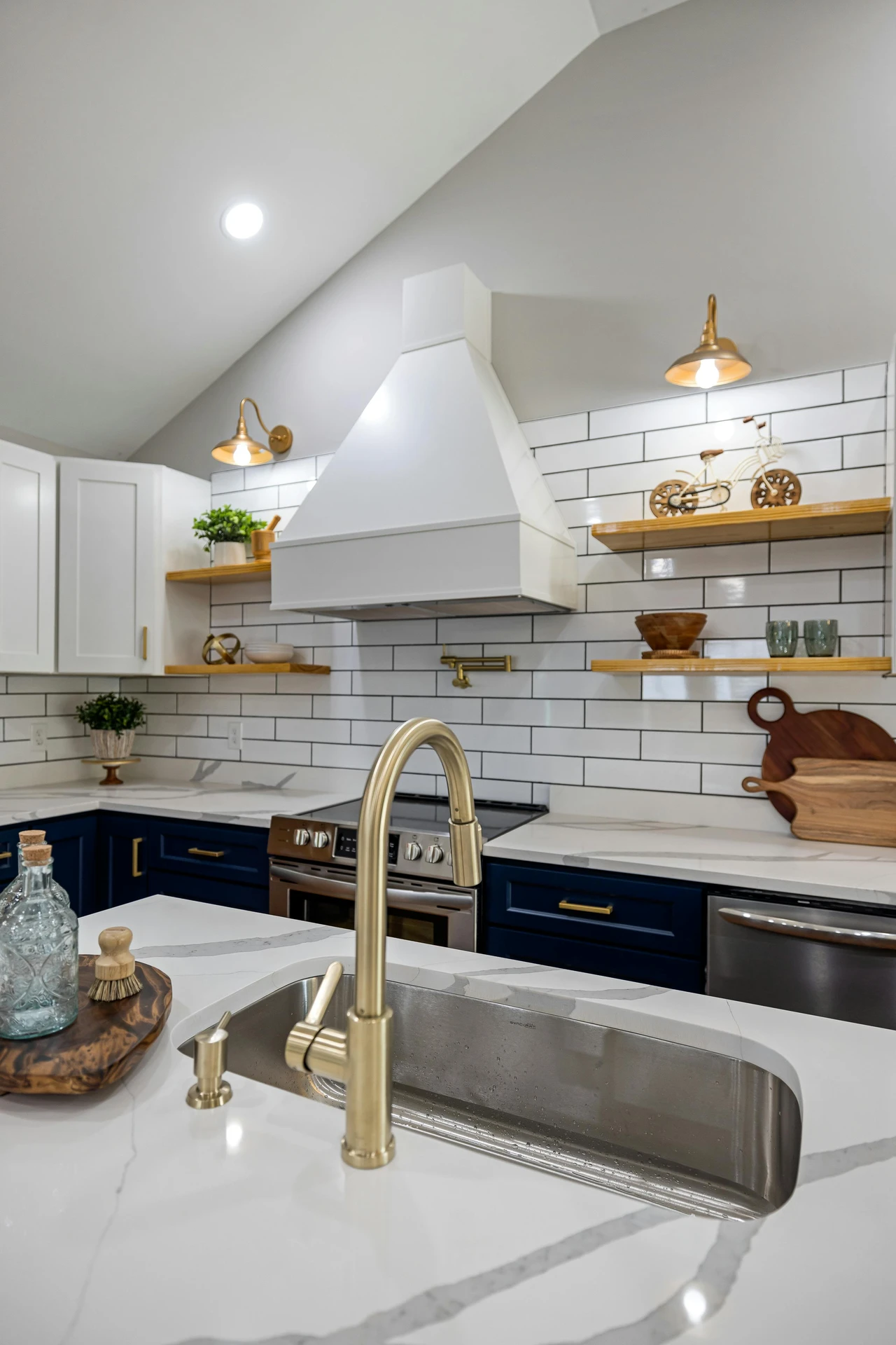 Charming and stylish kitchen featuring a white marble island with brass fixtures and a wooden tray displaying glass bottles and brushes-an inviting example of kitchen island decor ideas. The surrounding space blends modern and farmhouse elements with navy lower cabinets, white subway tiles, and open wooden shelves displaying plants, bowls, and playful accents, all contributing to a warm and cozy decor aesthetic.