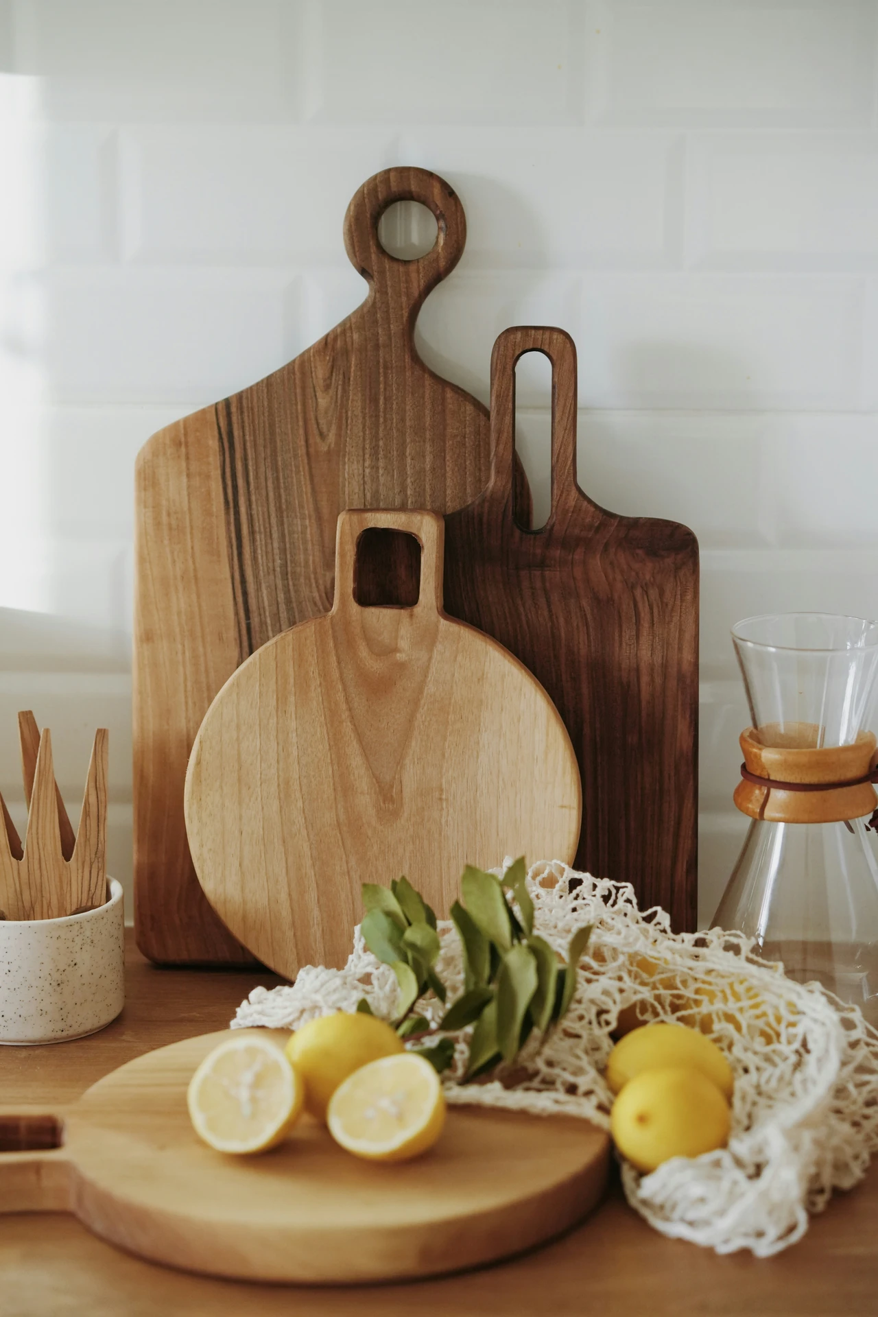Rustic kitchen scene showcasing a charming arrangement of wooden cutting boards in various shapes and tones, perfect for natural kitchen island decor ideas. Fresh lemons and greenery rest beside a mesh bag and glass carafe, adding a touch of warmth and organic style. This setup highlights how a simple cutting board display can elevate the look of a modern farmhouse or minimalist kitchen island.