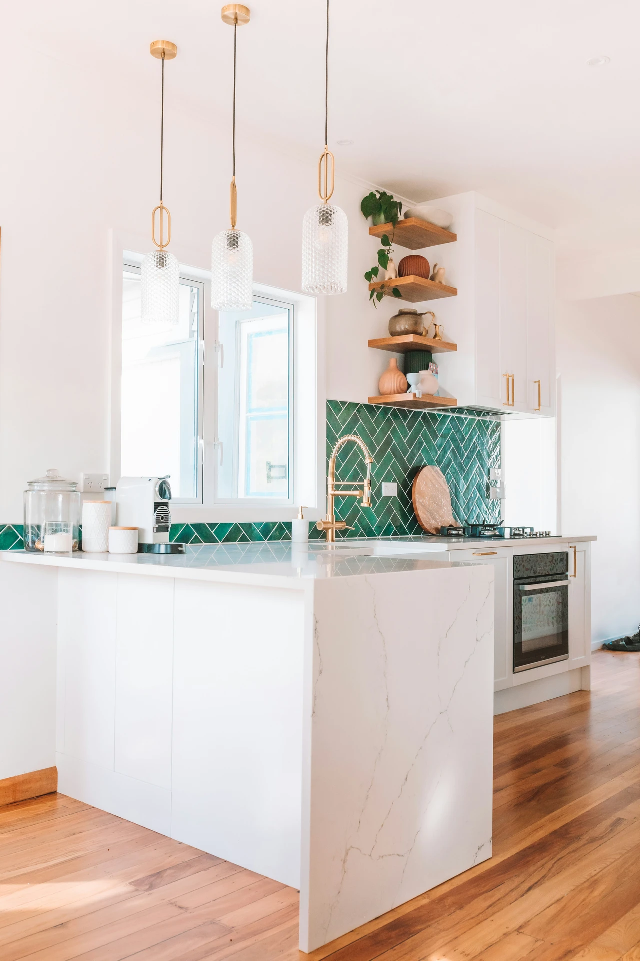 Bright and modern kitchen featuring a white marble kitchen island with minimalist decor, perfect for kitchen island decor ideas. The space includes a striking green tile backsplash in a herringbone pattern, gold hardware and faucet, and warm wood accents. Floating shelves display a mix of ceramics and greenery, adding charm and functionality.