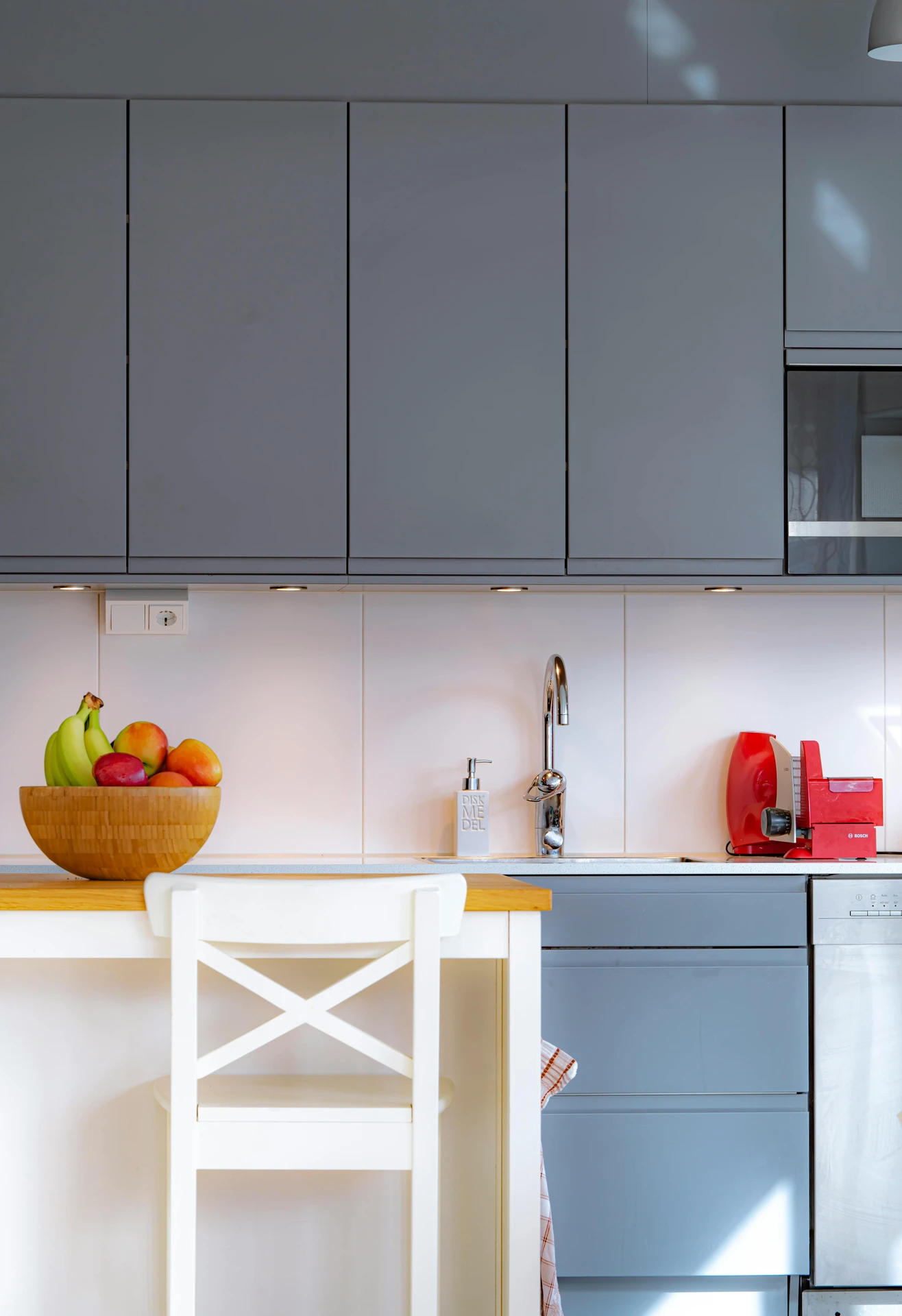 Bright and functional kitchen featuring sleek gray cabinets and a minimalist white island with wooden countertop accents. A vibrant fruit bowl filled with bananas and apples adds a pop of color, showcasing simple yet effective kitchen island decor ideas. The scene is completed with a red toaster and coffee machine, blending modern practicality with a touch of personality.