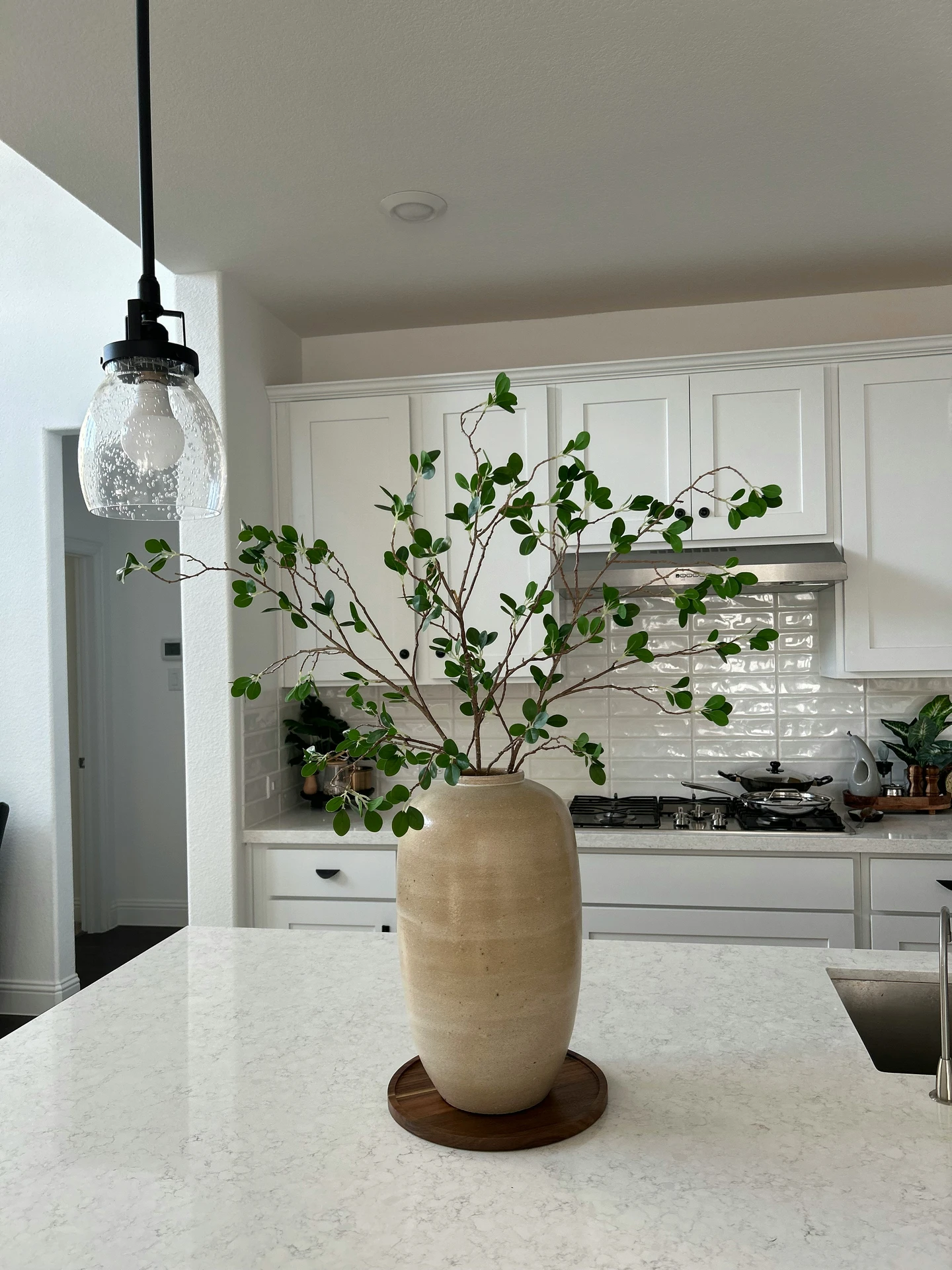 Bright and airy kitchen featuring a minimalist white quartz island adorned with a tall ceramic vase filled with greenery, offering a fresh and natural touch. The arrangement serves as a stunning centerpiece and exemplifies simple yet elegant kitchen island decor ideas that bring life and movement to the space. Perfect for a clean, modern home with organic accents.
