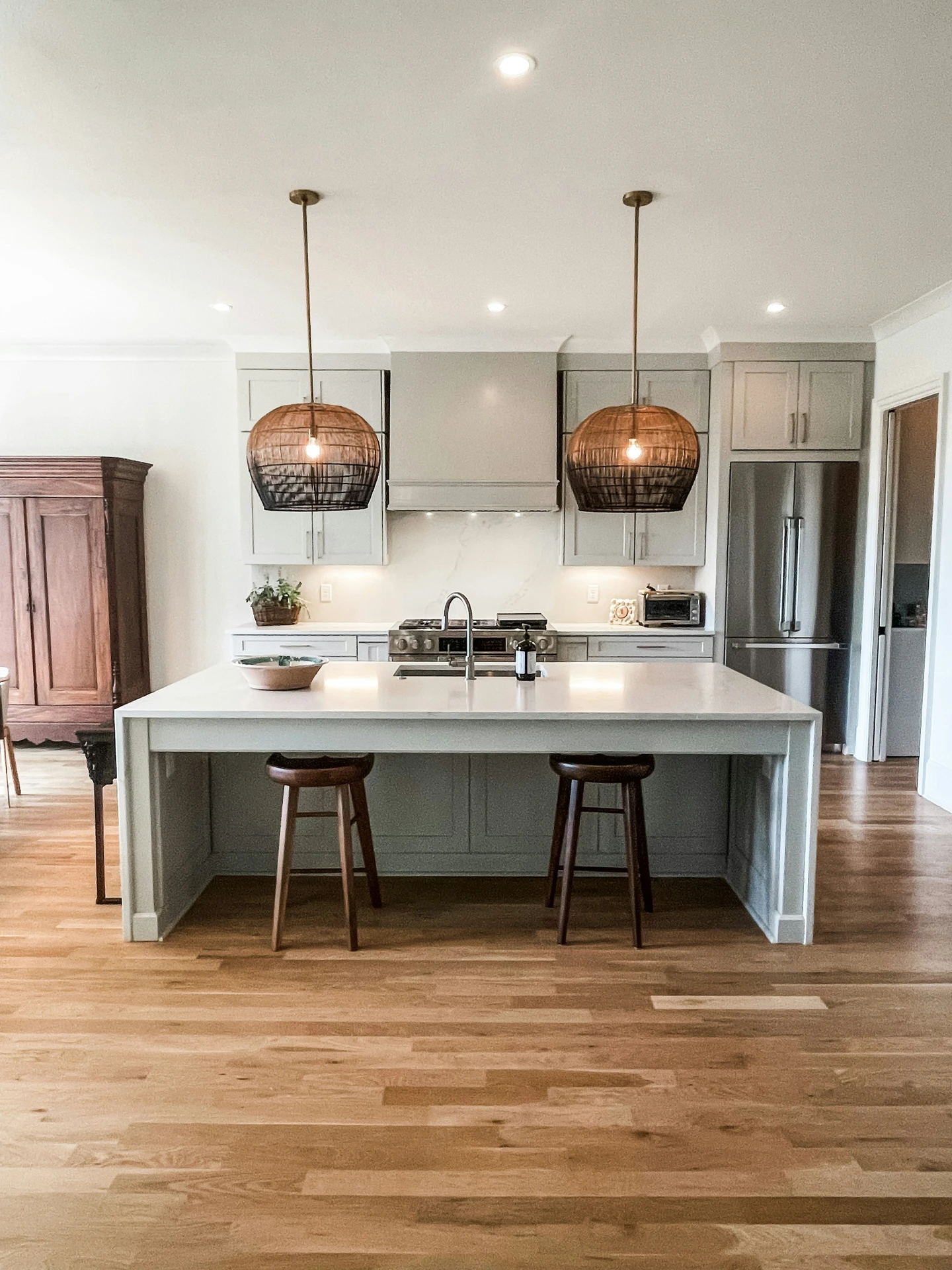 Spacious modern kitchen with a large central island featuring subtle kitchen island decor ideas like a simple bowl and potted plant, creating a clean yet inviting atmosphere. Two woven pendant lamps serve as stylish hanging lights above the kitchen island, adding warmth and texture to the neutral-toned space with wood flooring and light gray cabinetry.