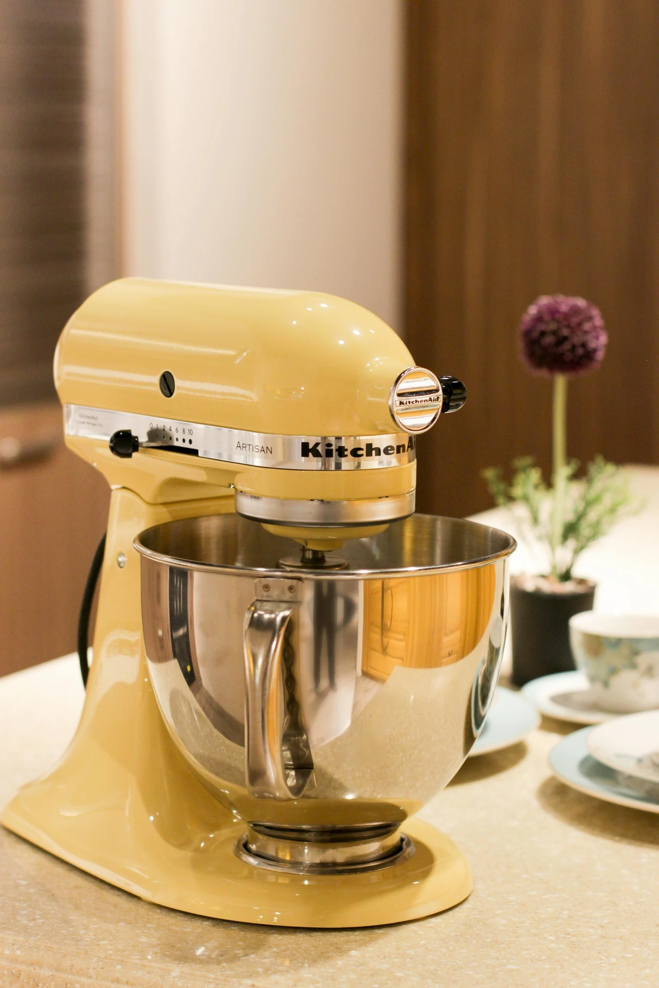 Close-up of a yellow KitchenAid stand mixer placed on a light stone countertop, accompanied by delicate teacups and a small potted plant in the background. This setup offers charming kitchen island decor ideas, showing how to display a kitchen machine in a way that blends functionality with visual appeal in a cozy and stylish kitchen setting.